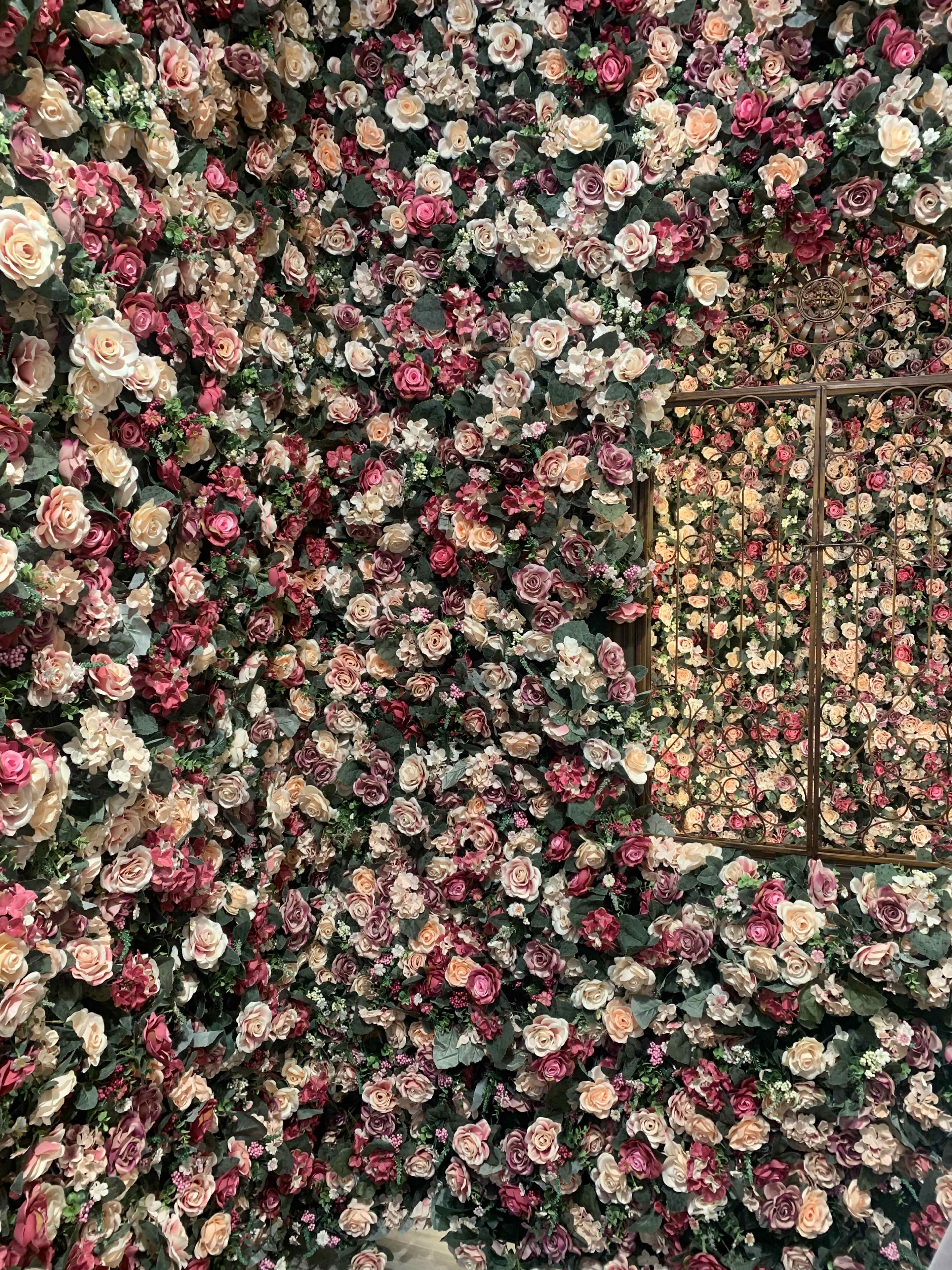 The scene features a room covered in a dense arrangement of artificial flowers, with a mirror reflecting the floral decor.