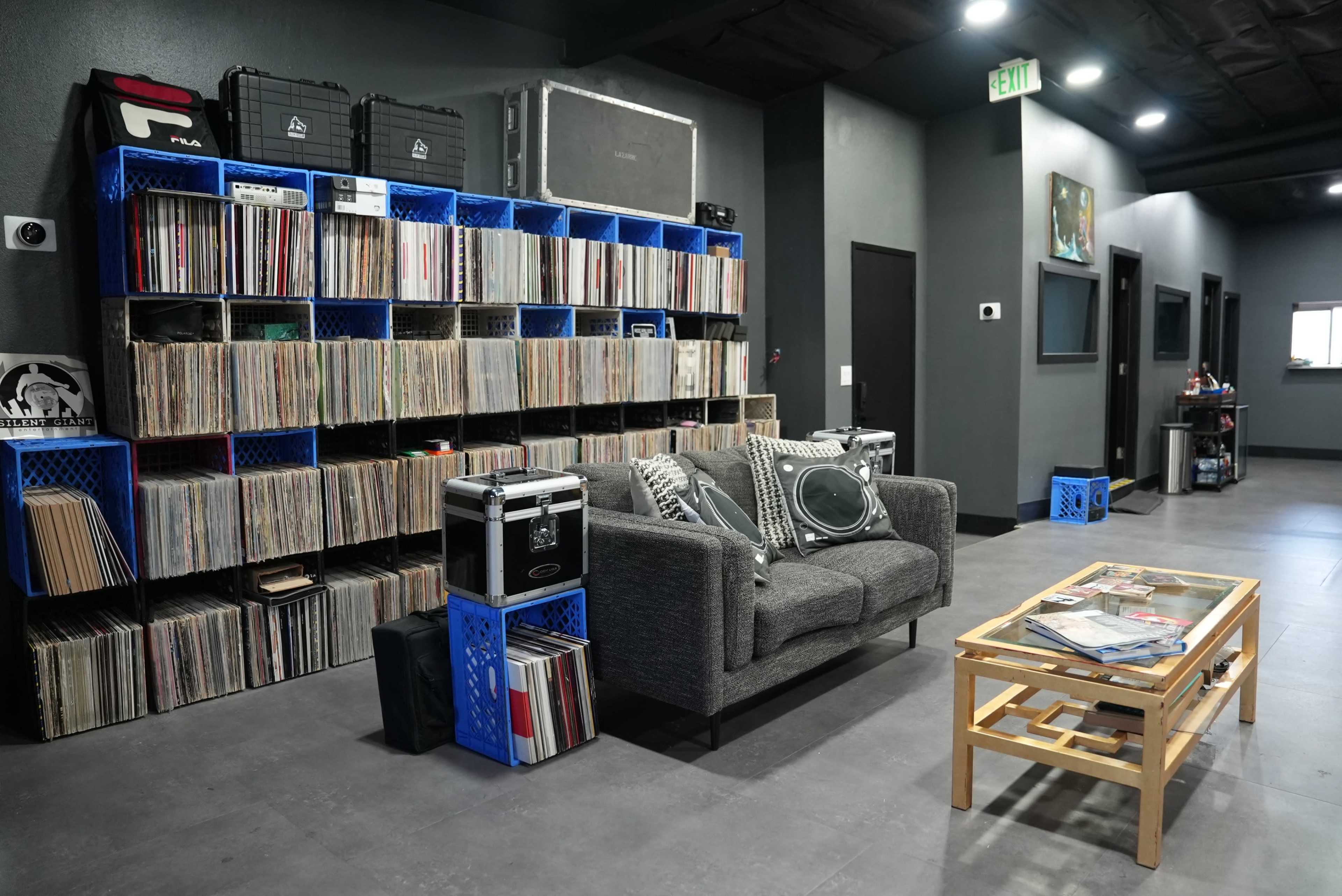The image shows a modern room with a wall of neatly organized vinyl records and a gray couch next to a coffee table stacked with magazines.