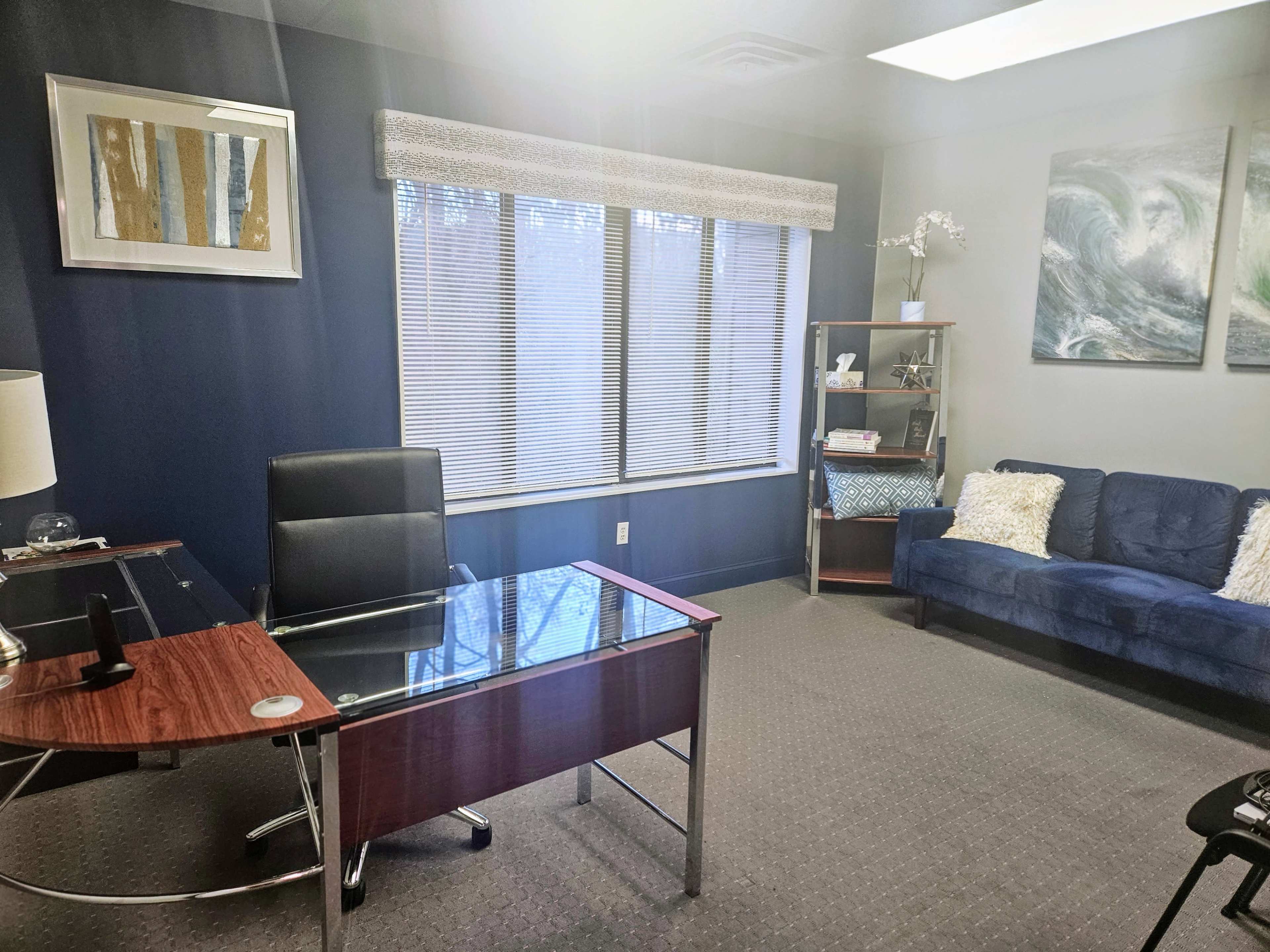 An office space featuring a glass-top desk, a black office chair, a blue couch, a bookshelf, and a large window with blinds.