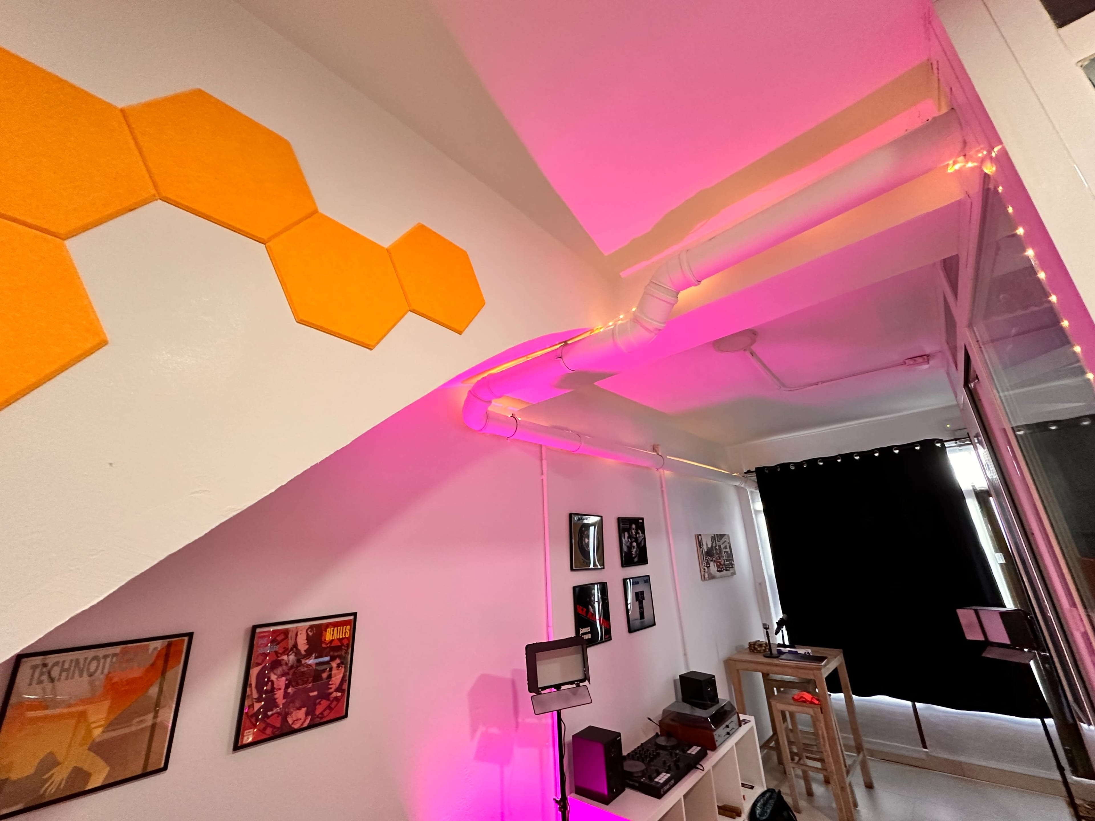 The image shows a room with orange hexagonal wall decorations, pink LED lights along the ceiling, and a workspace featuring a computer setup and framed posters.