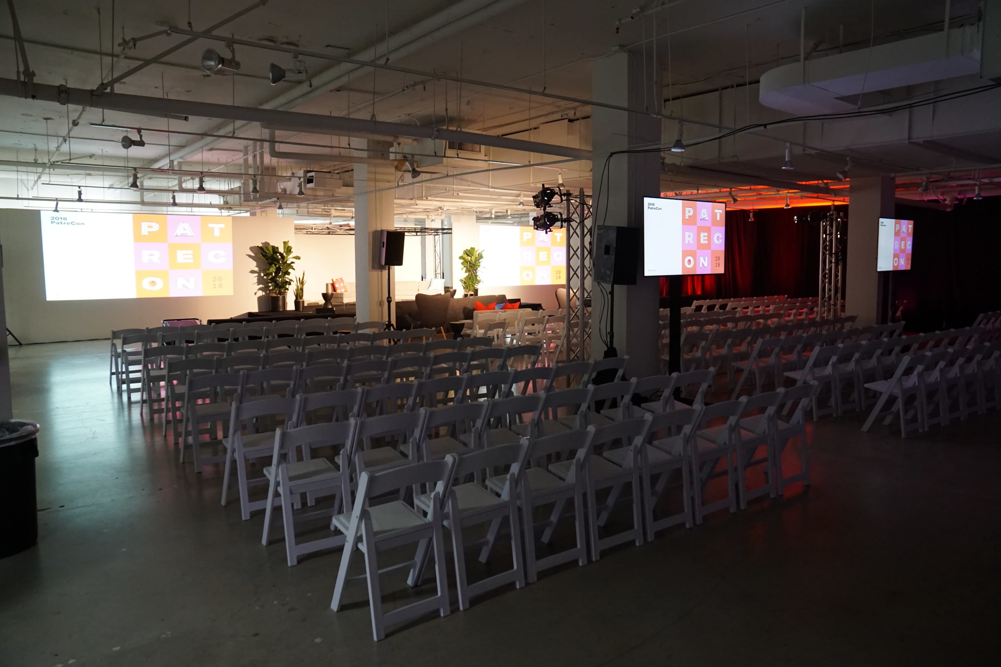 The image shows an event space with rows of white chairs arranged for attendees, a stage set up with screens, and plants placed around the room.