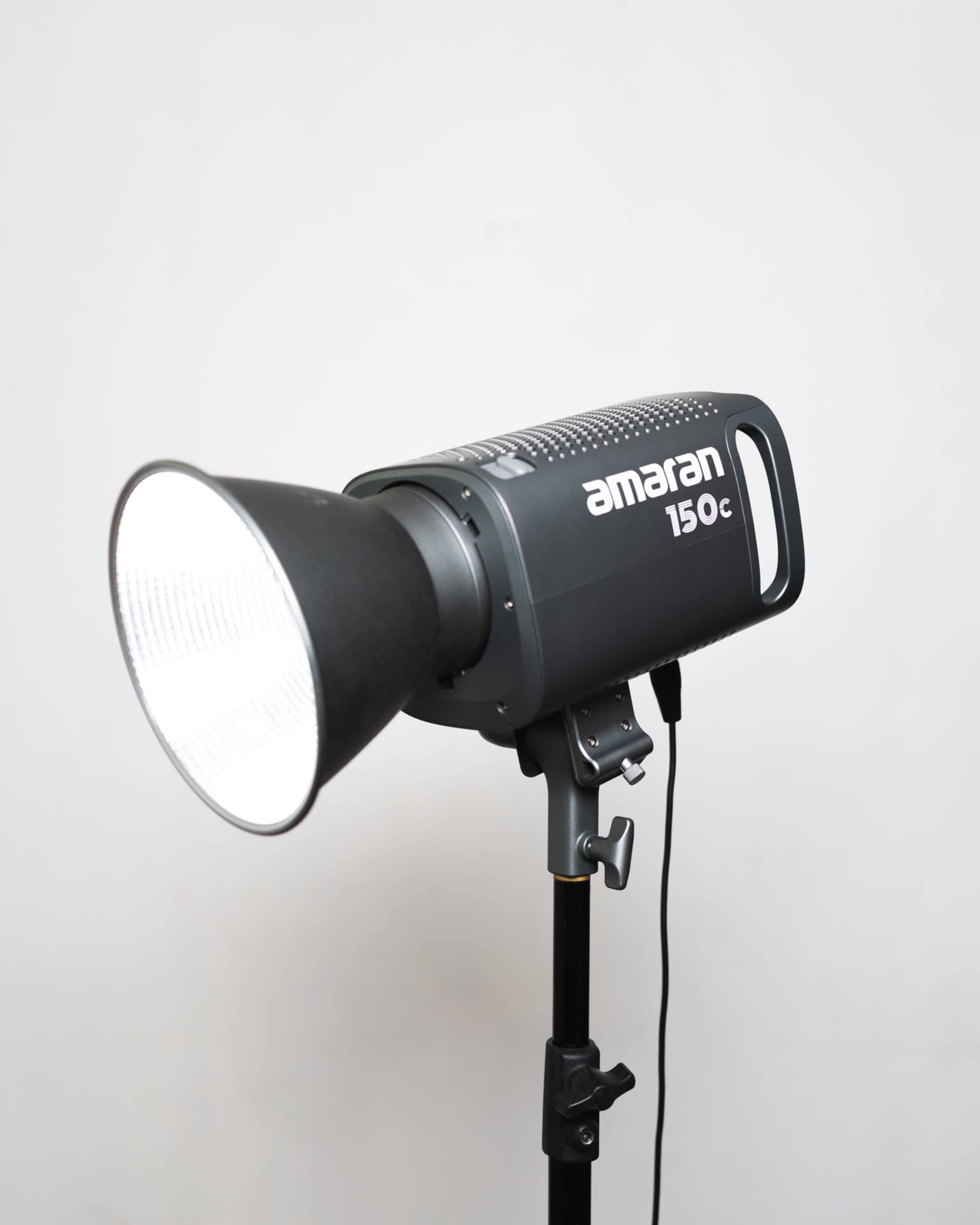 A studio lighting device mounted on a stand, specifically an Amaran 150c with a reflector attached.