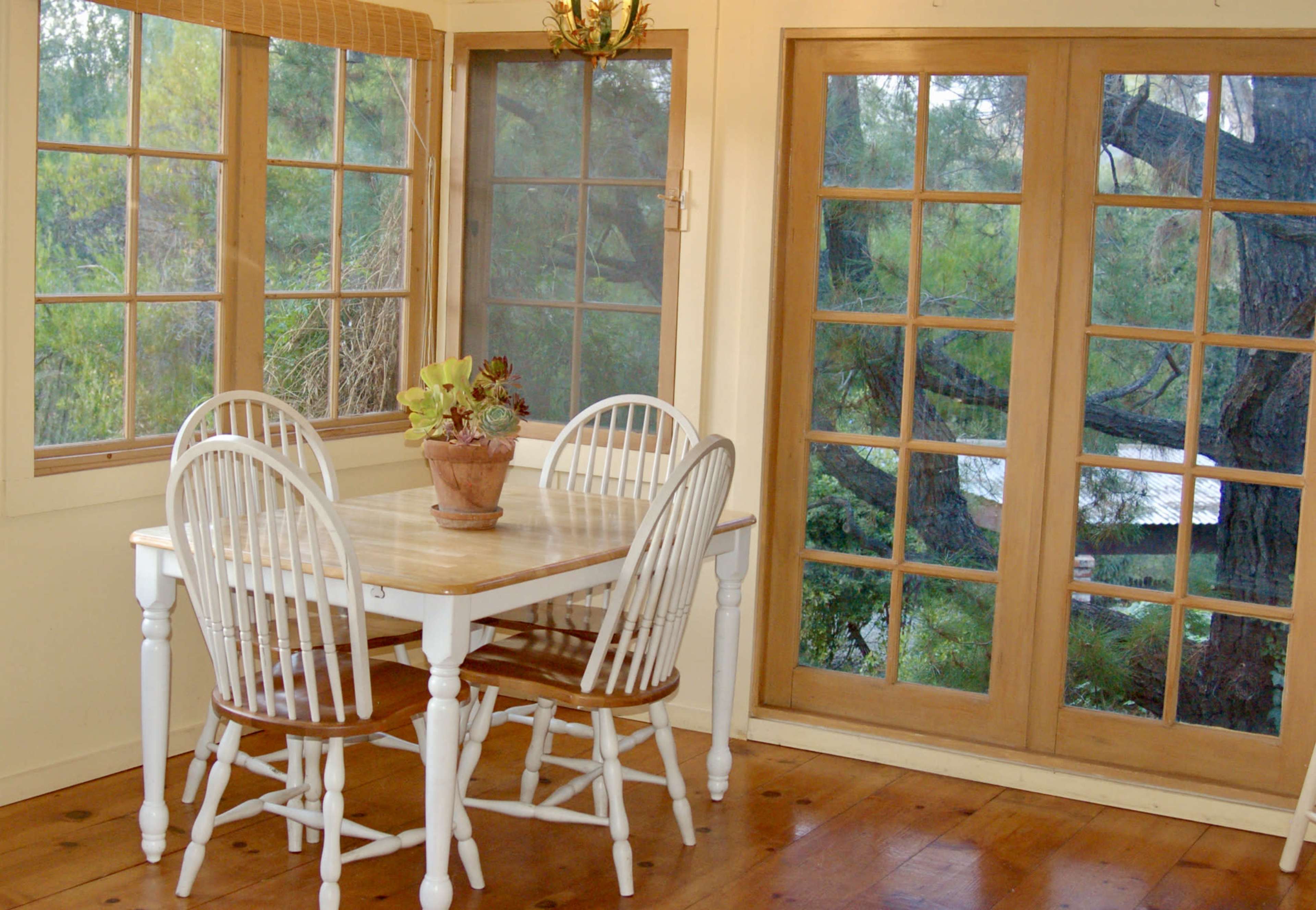 A wooden table surrounded by four chairs sits in a sunlit room with large windows overlooking a natural scene.