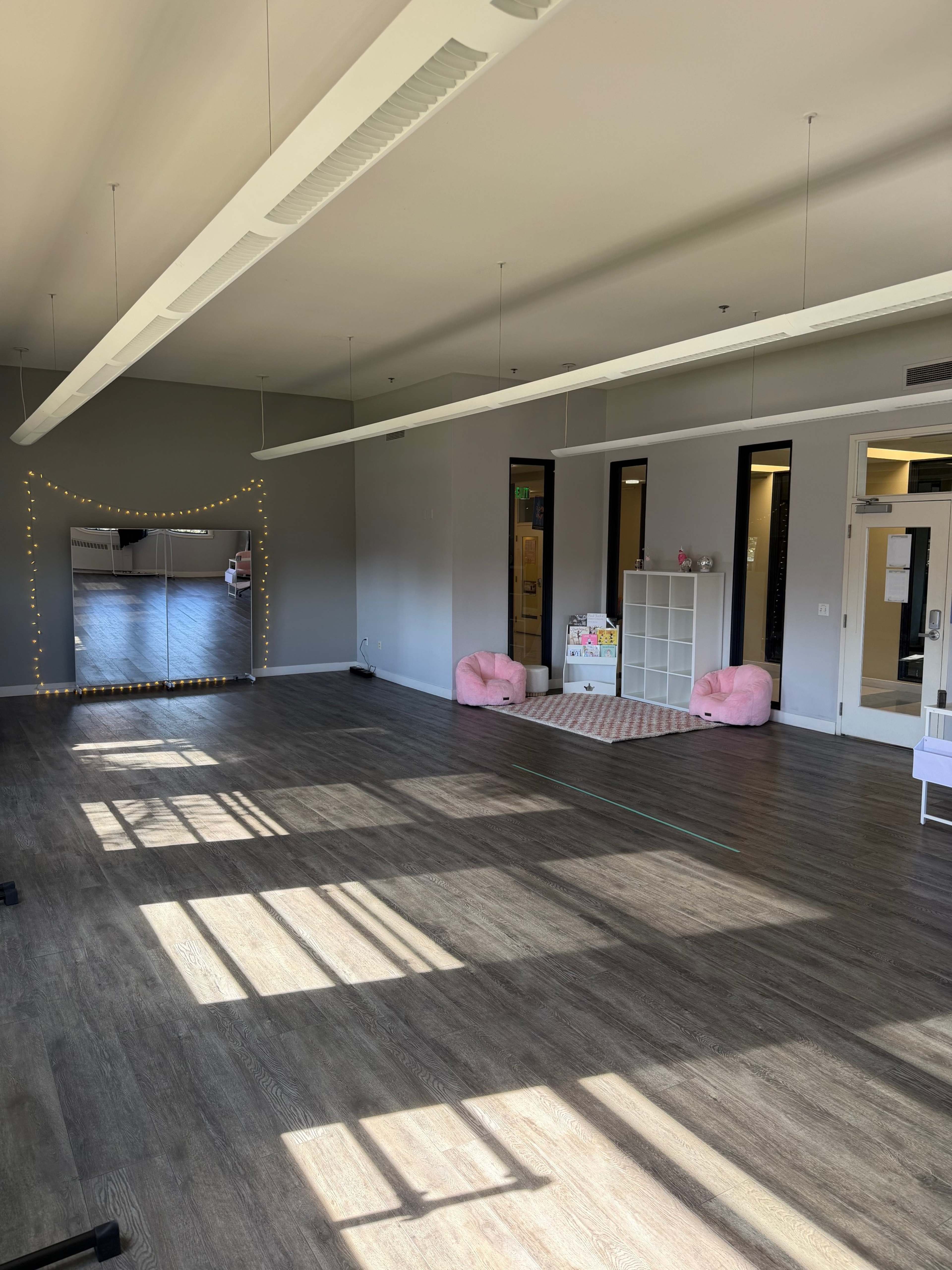 The image shows a spacious, well-lit room with wooden floors, a large mirror on one wall, and two pink seating areas at one end.