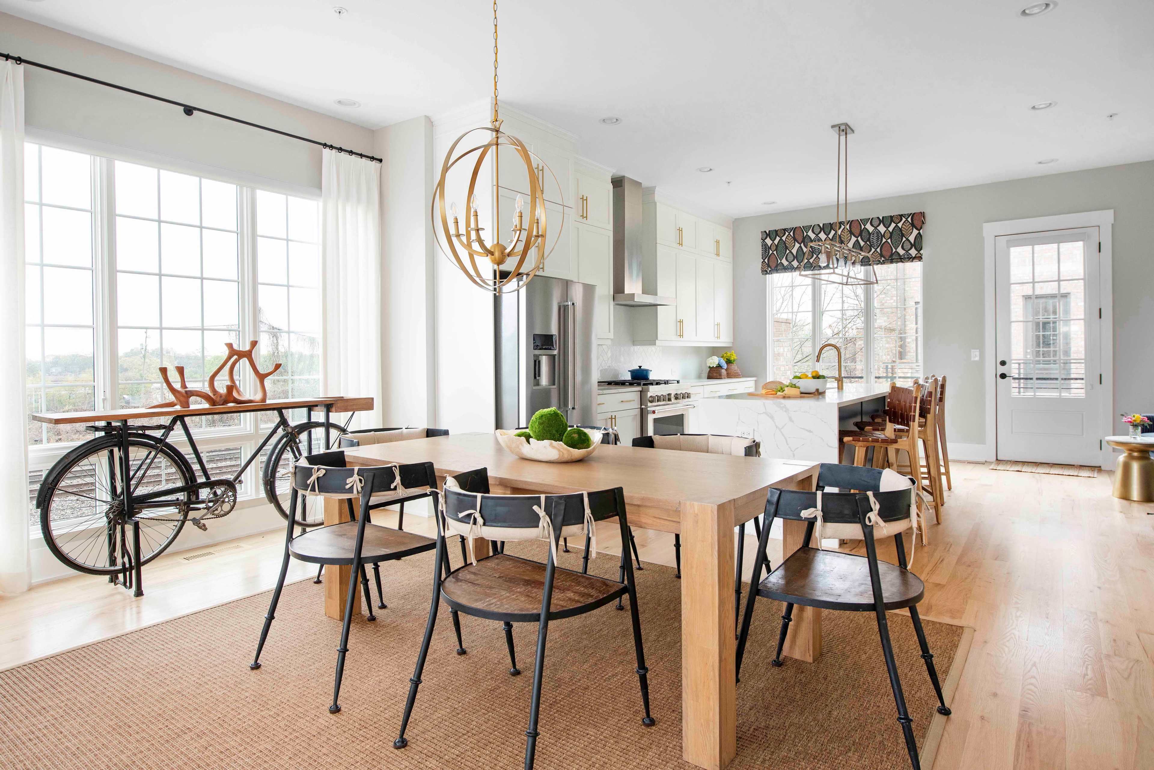 The image shows a modern kitchen and dining area with a large wooden table surrounded by black chairs, a bicycle used as decor, and large windows allowing natural light to fill the space.