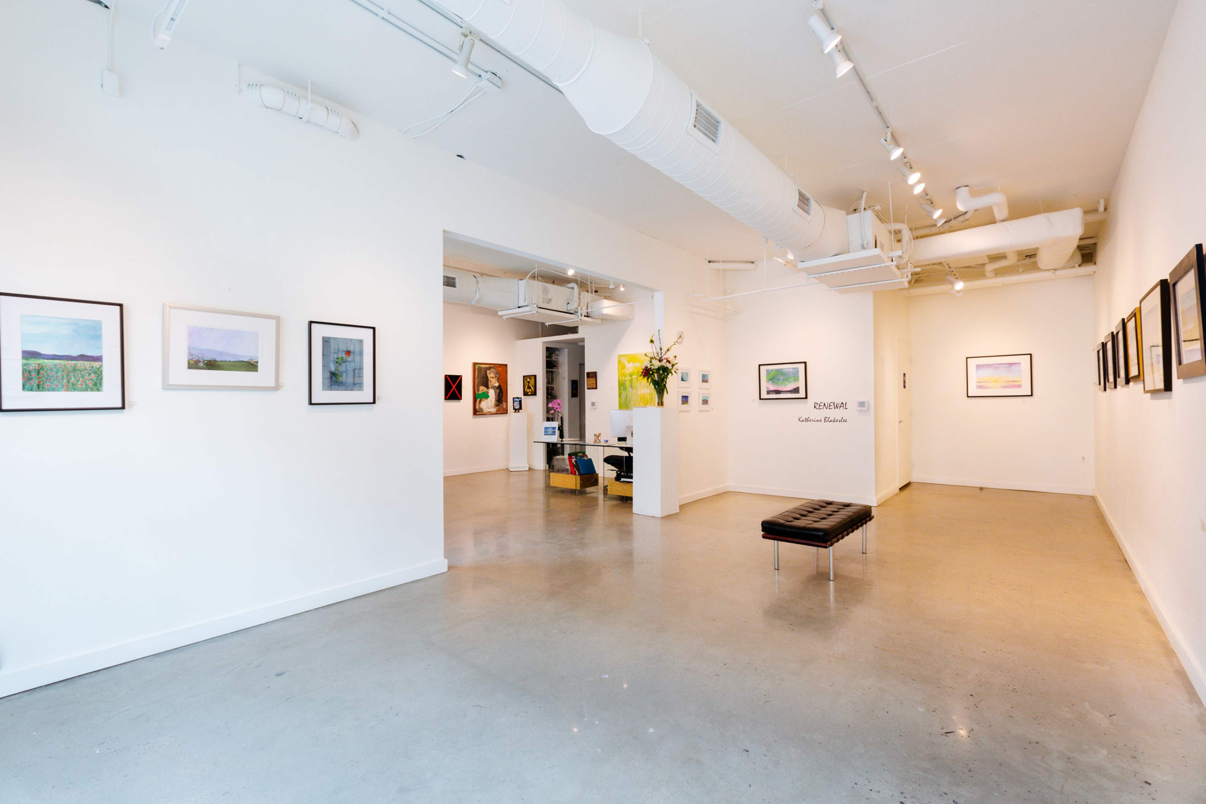 The image shows a spacious art gallery with white walls, featuring framed artworks displayed on the walls and a bench in the foreground.