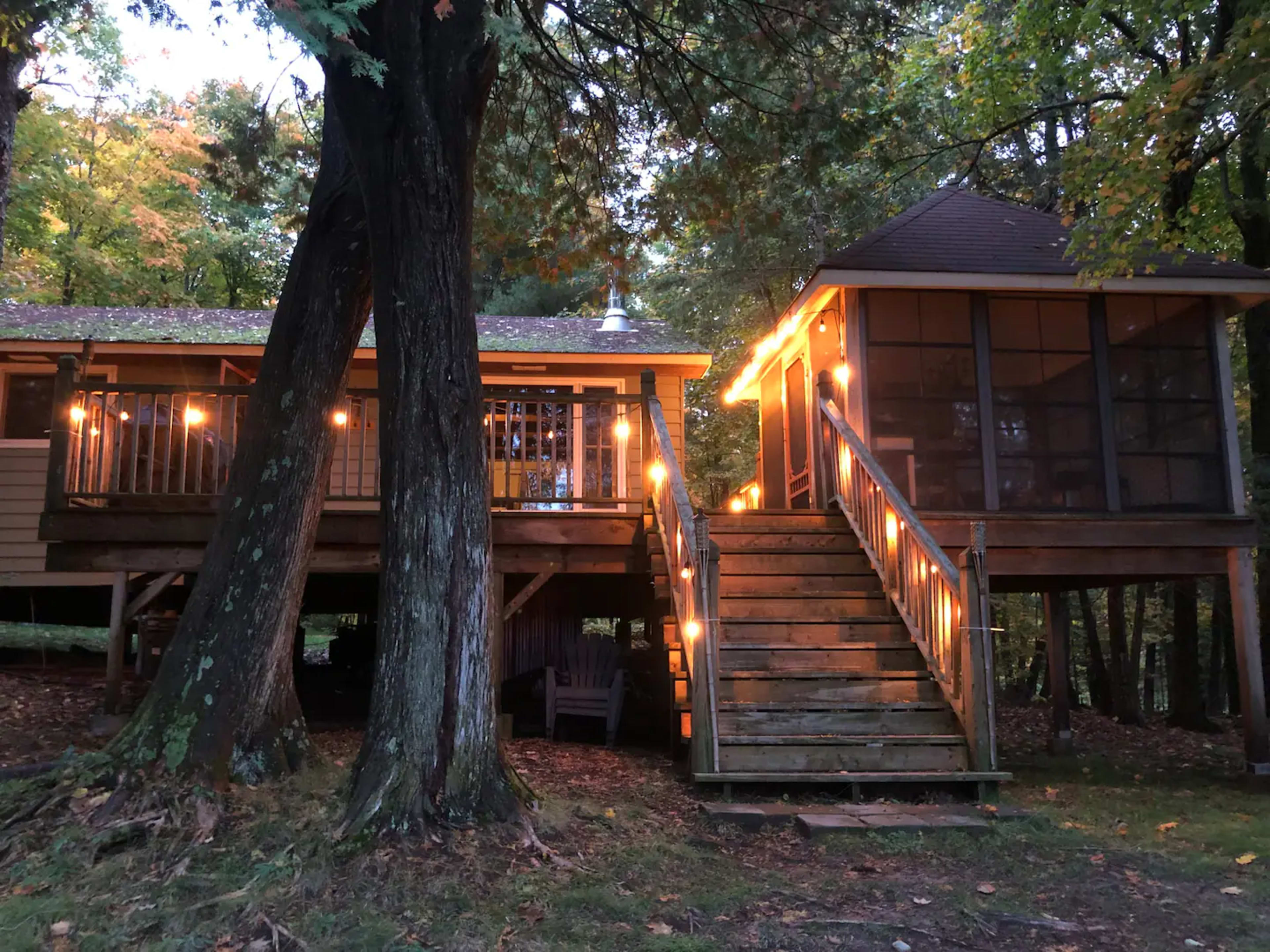 A wooden cabin and a screened gazebo are connected by stairs and adorned with string lights in a wooded area.