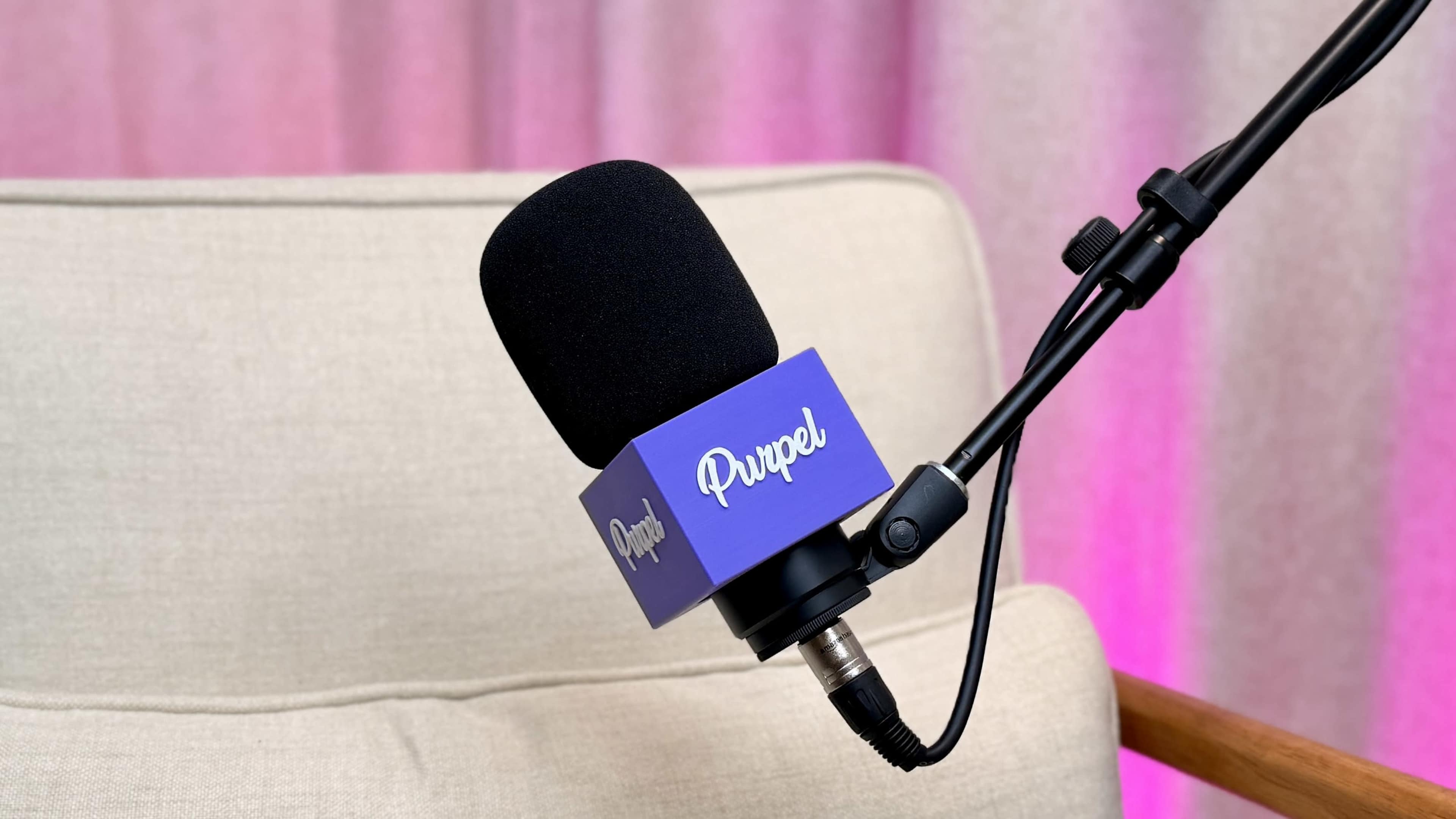 A black microphone with a purple logo is mounted on a chair's armrest against a softly lit pink background.
