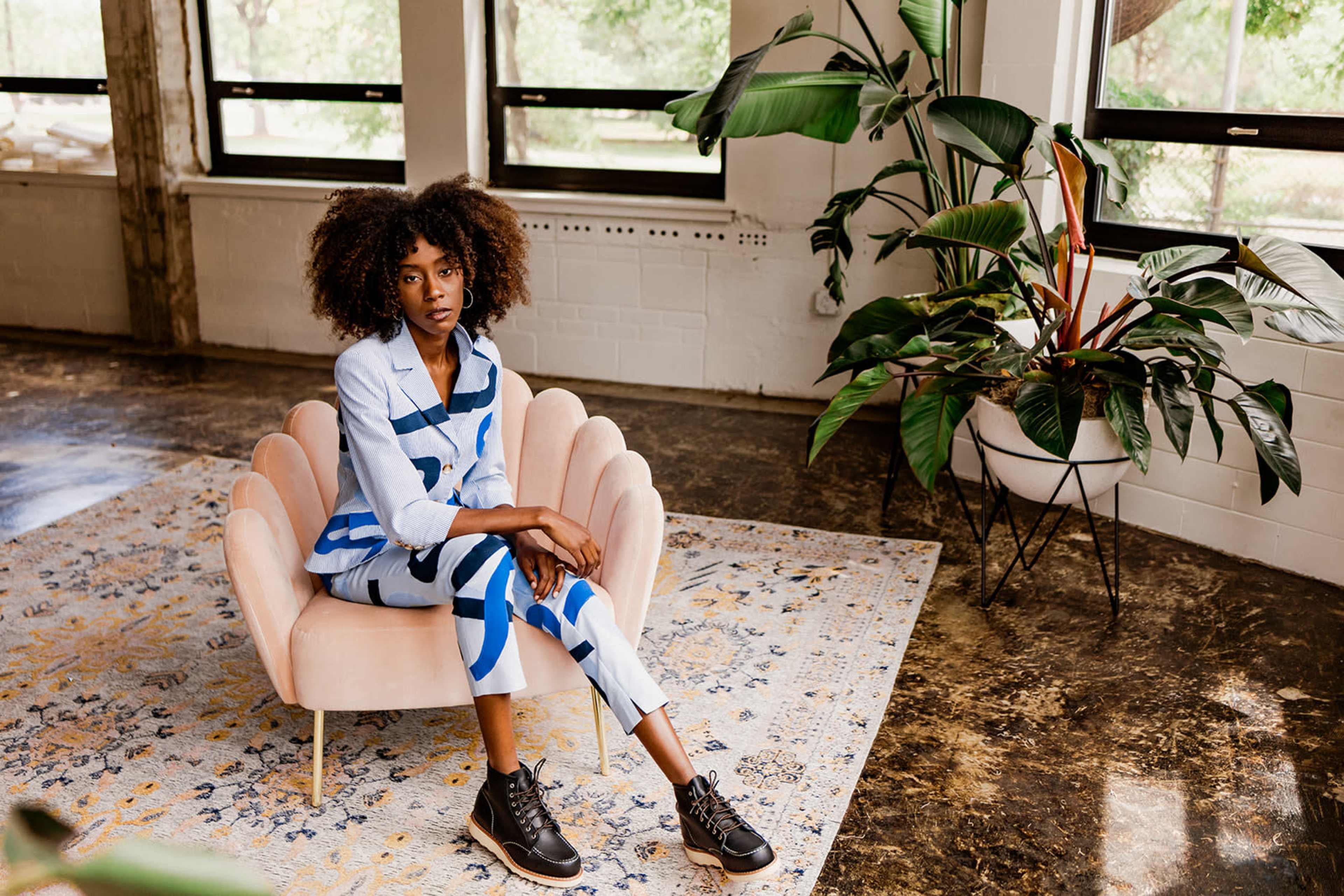 A person sits on a pink chair in a modern indoor space decorated with plants and a patterned rug.