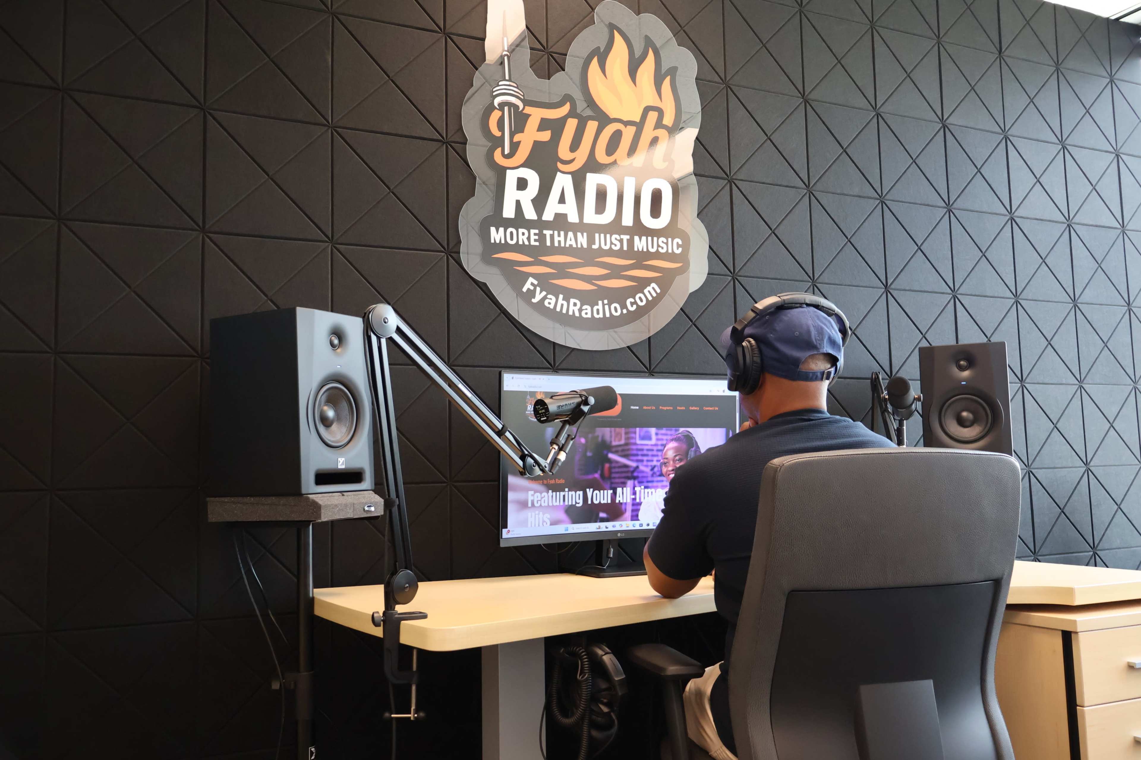 A person sits at a desk with headphones on, focused on a computer screen in a stylish radio studio adorned with promotional branding.