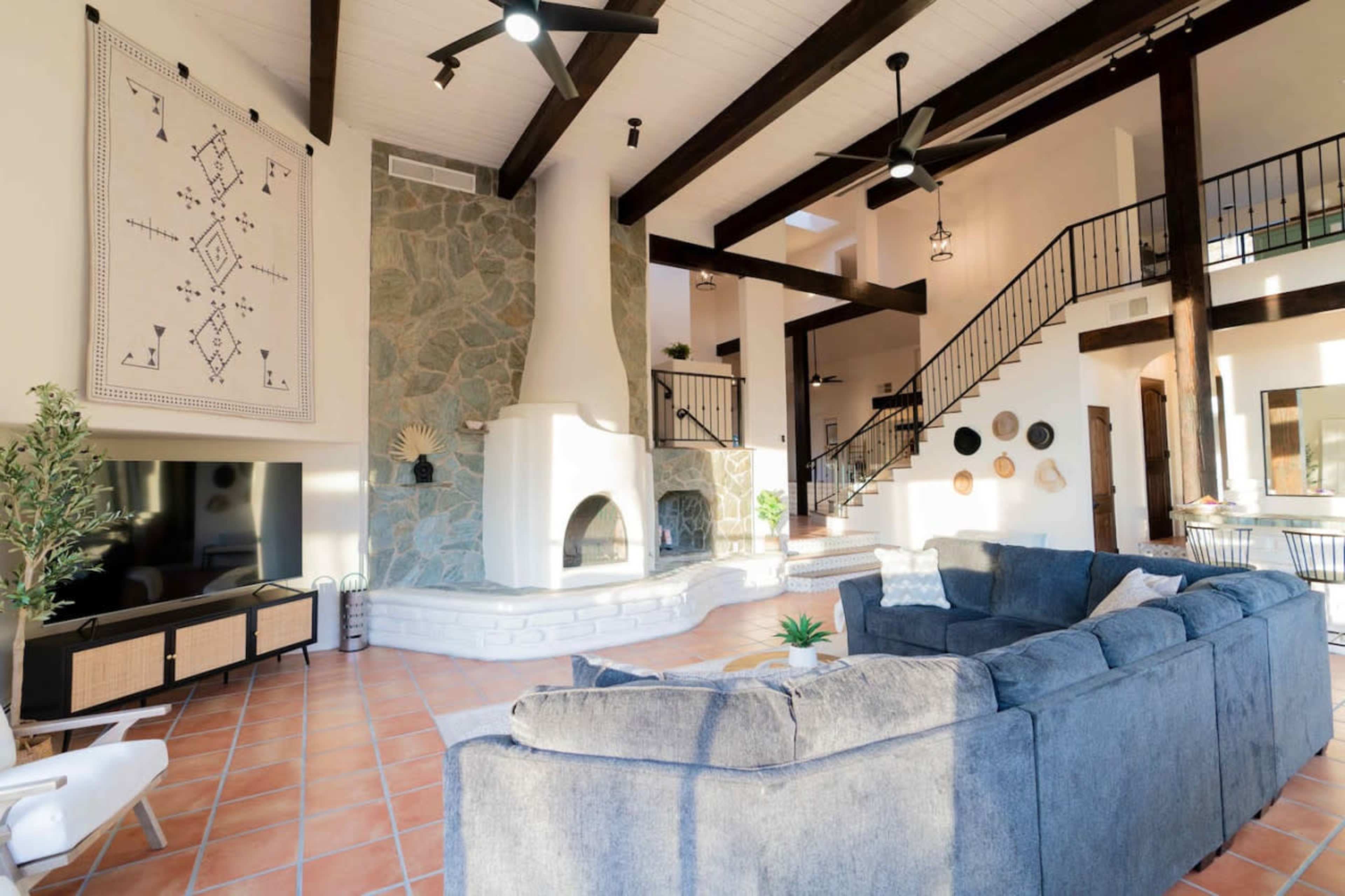 The living room features a large blue sectional sofa, a stone fireplace, and high wooden beams, with natural light streaming through large windows.