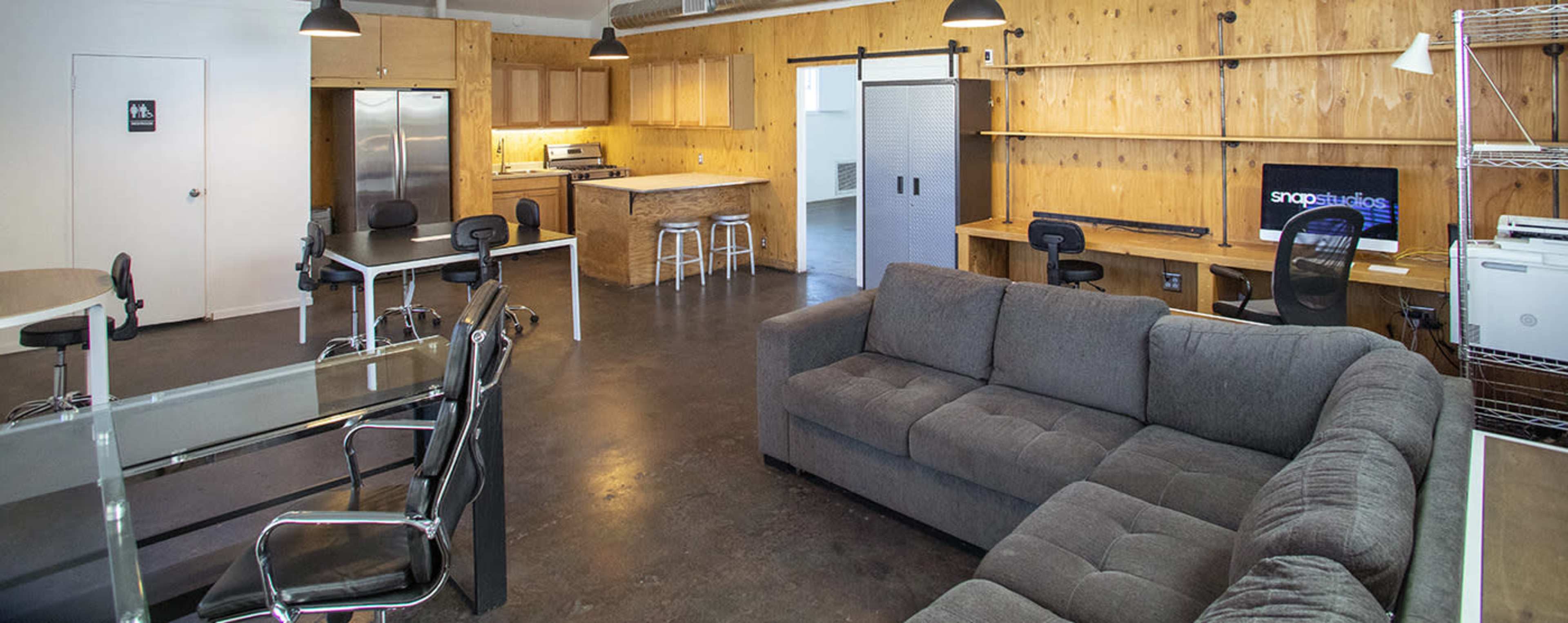 A modern office space with a gray sectional sofa, a glass table, several chairs, a kitchenette area, and workstations along wooden walls.