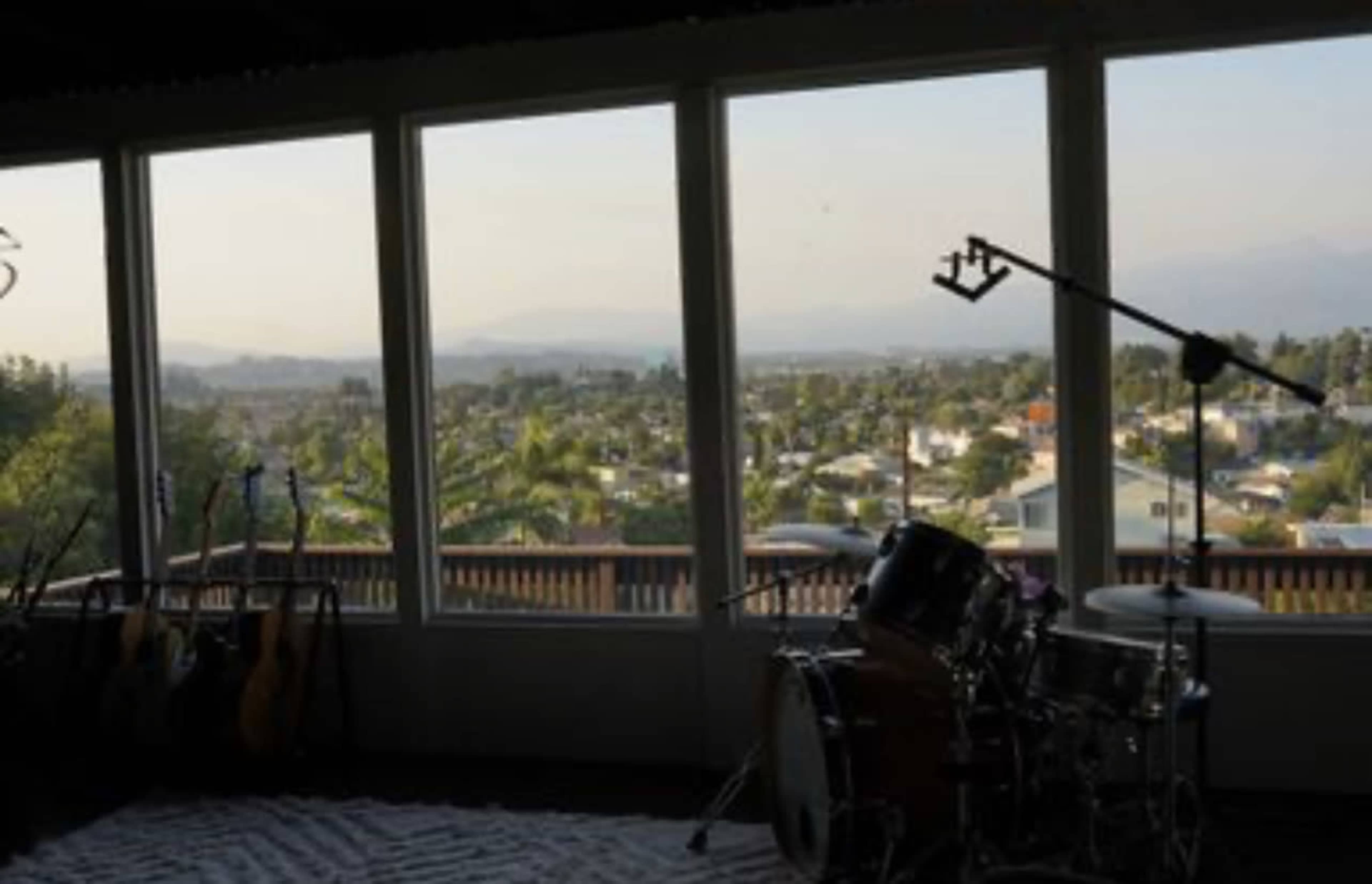The image shows a room with large windows overlooking a residential area and mountains in the distance, with musical instruments, including guitars and a drum set, positioned in the corner.