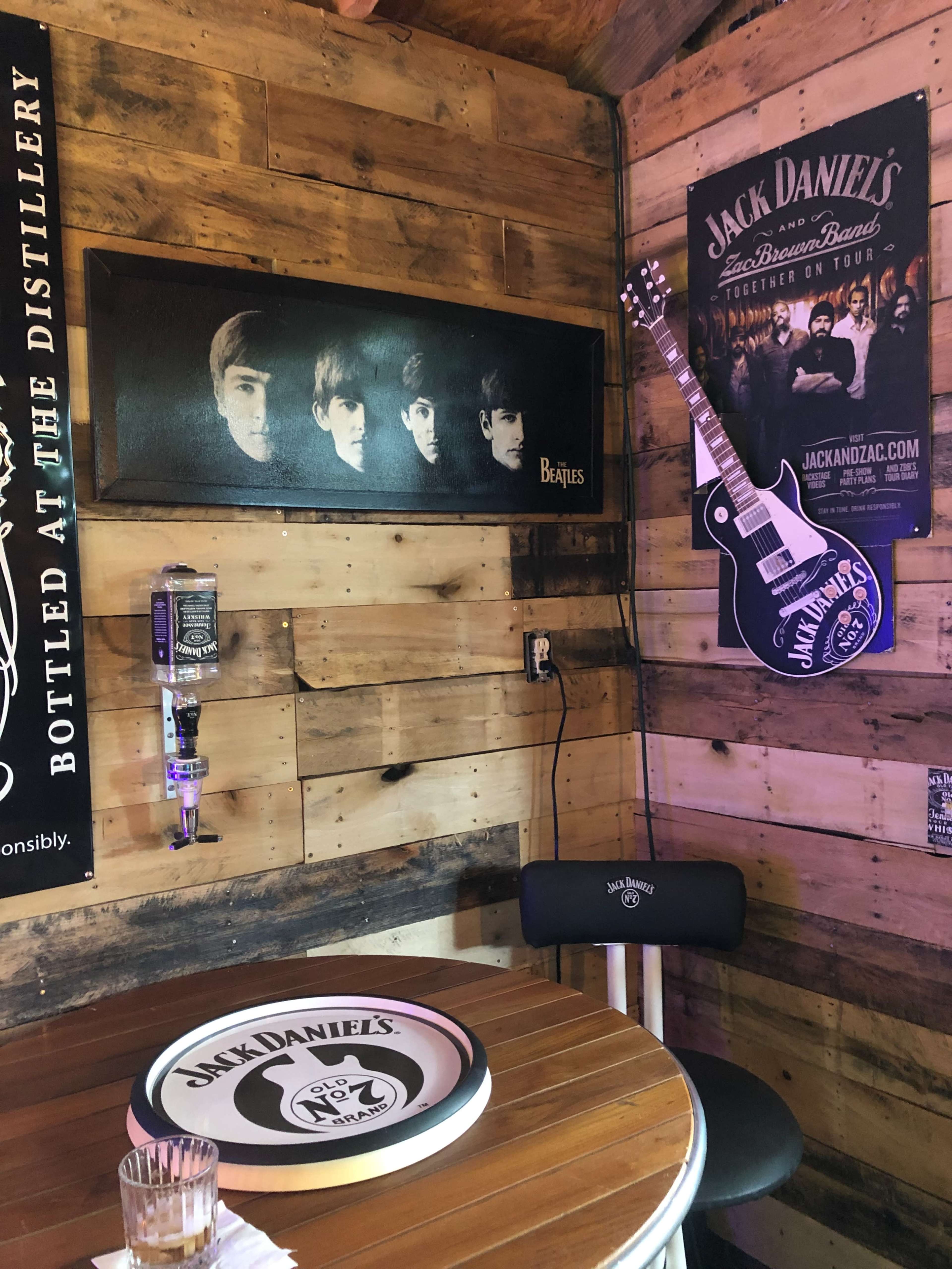 A rustic bar setting featuring a framed portrait of The Beatles on the wall, a Jack Daniel's guitar-shaped sign, and a round table with a Jack Daniel's logo.