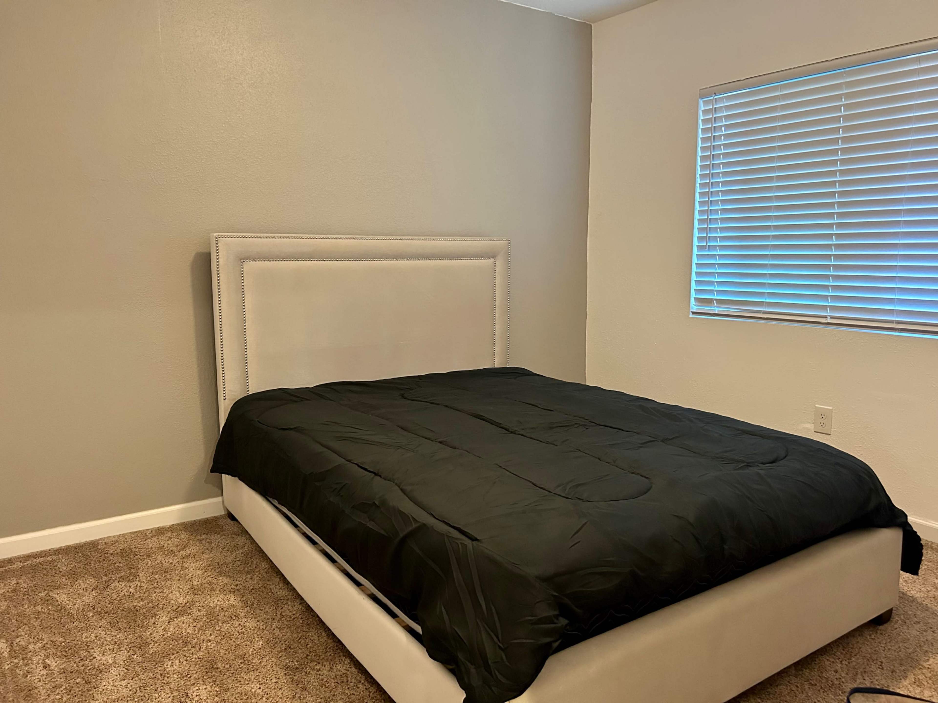 A bedroom with a black bedspread on a light-colored upholstered bed and a window with blinds.