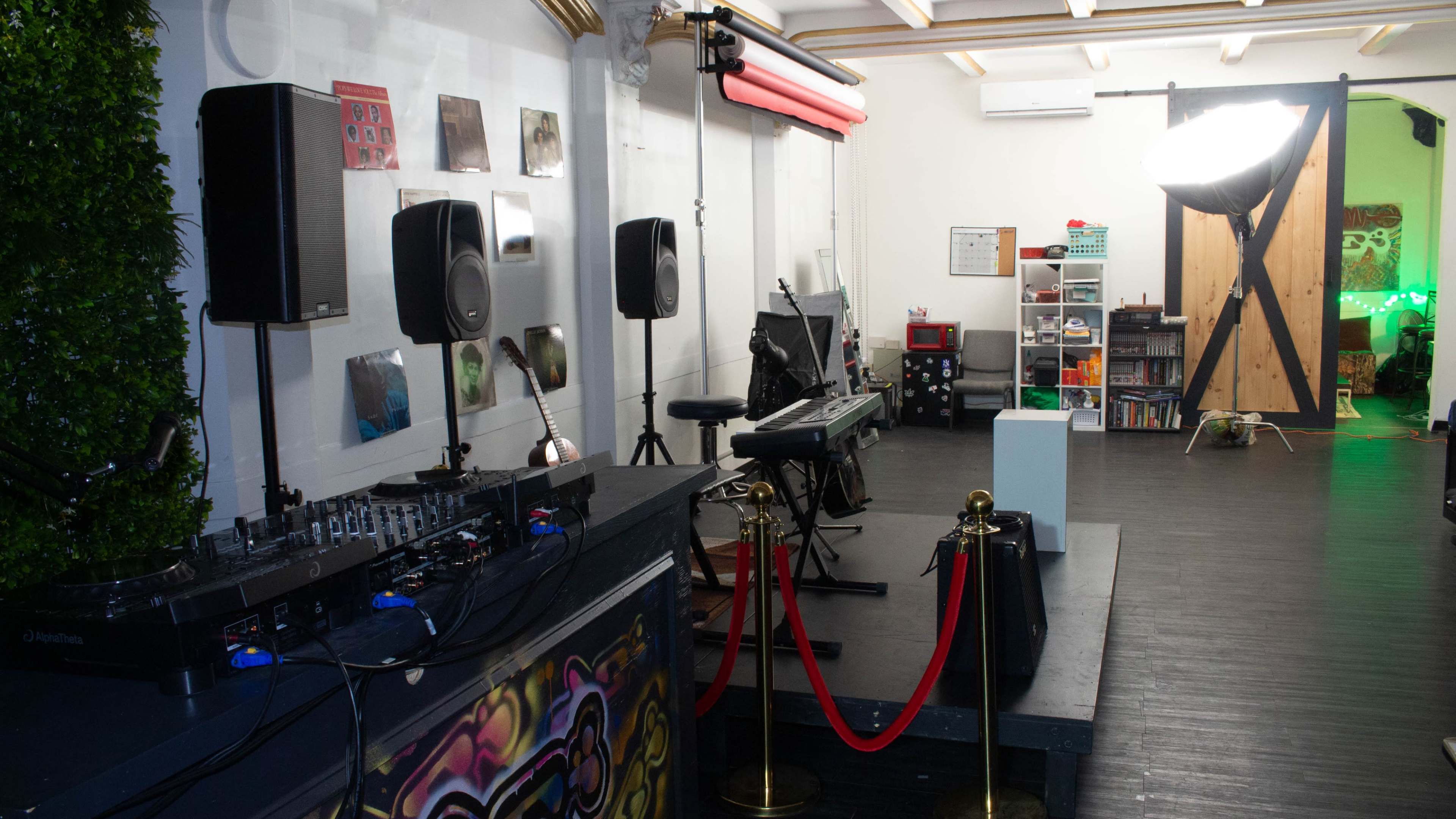 The image shows a music performance space featuring a DJ setup on the left, a keyboard and microphone on a small stage, and sound equipment positioned around the room.