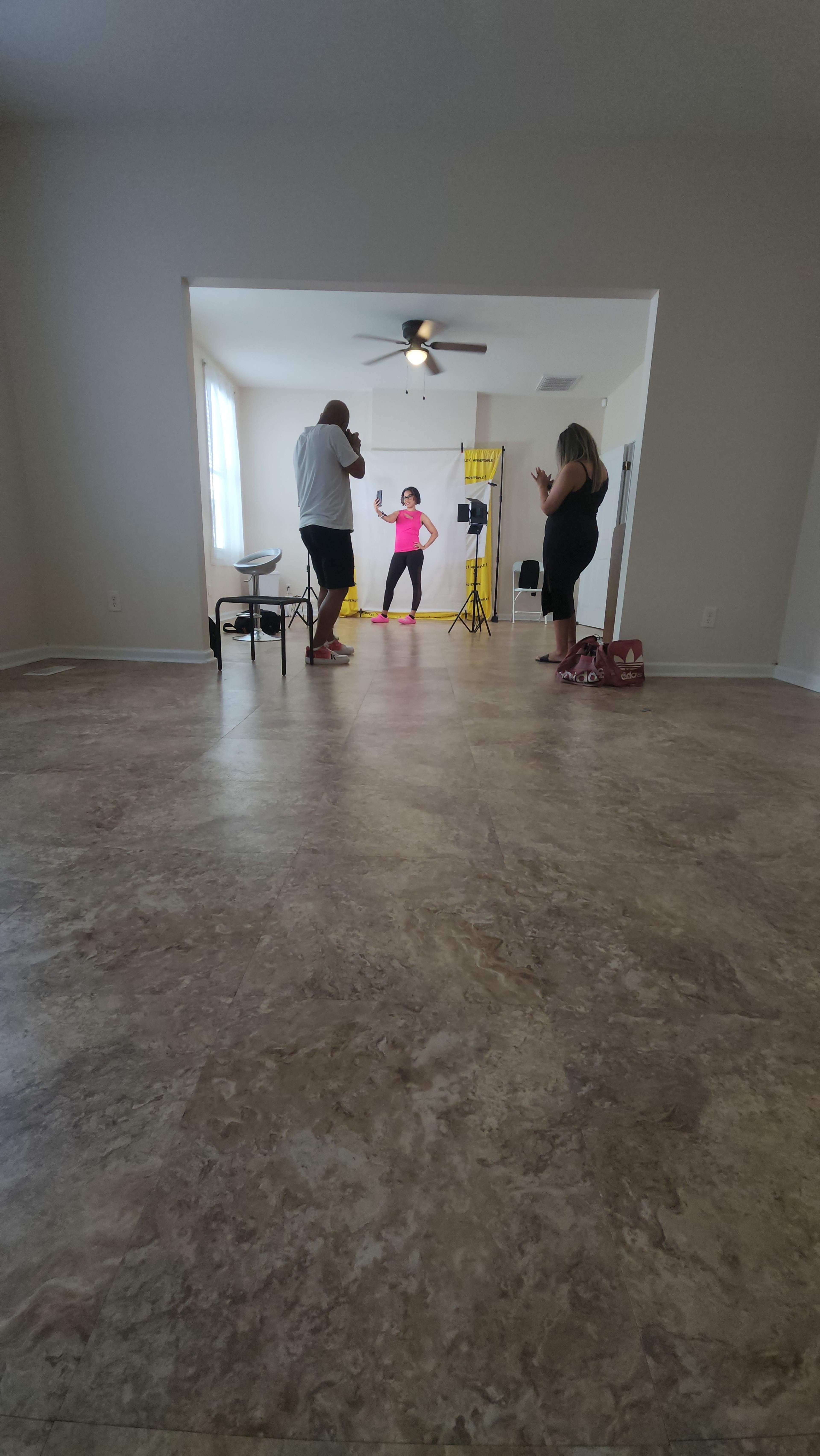A group of three people is participating in a photo shoot in a spacious, empty room with light-colored walls and a ceiling fan.