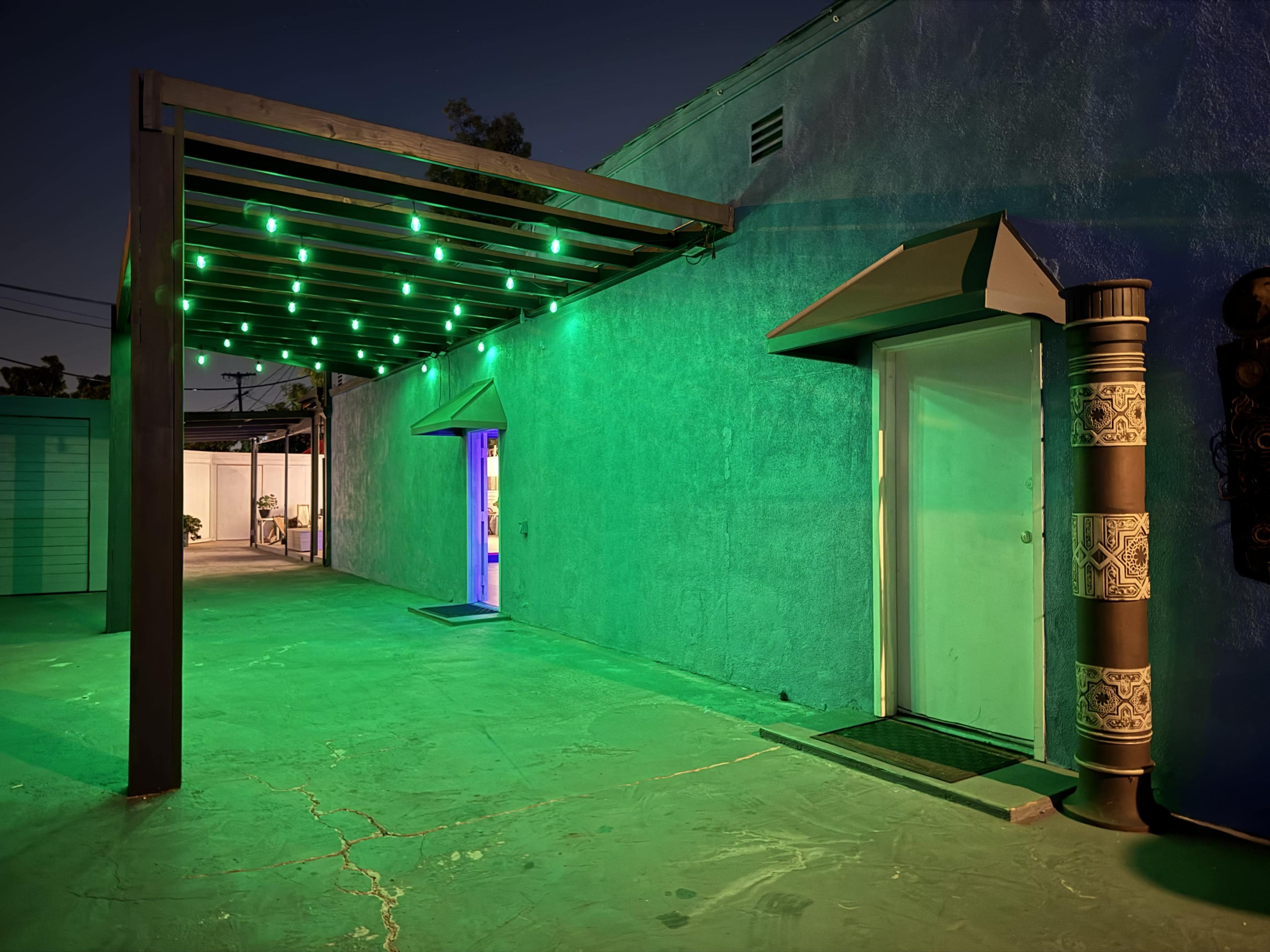 The scene shows a modern entrance with a green-lit overhang, leading to a door on the left and a contrasting blue-lit entrance on the right, set against a textured wall in an evening setting.