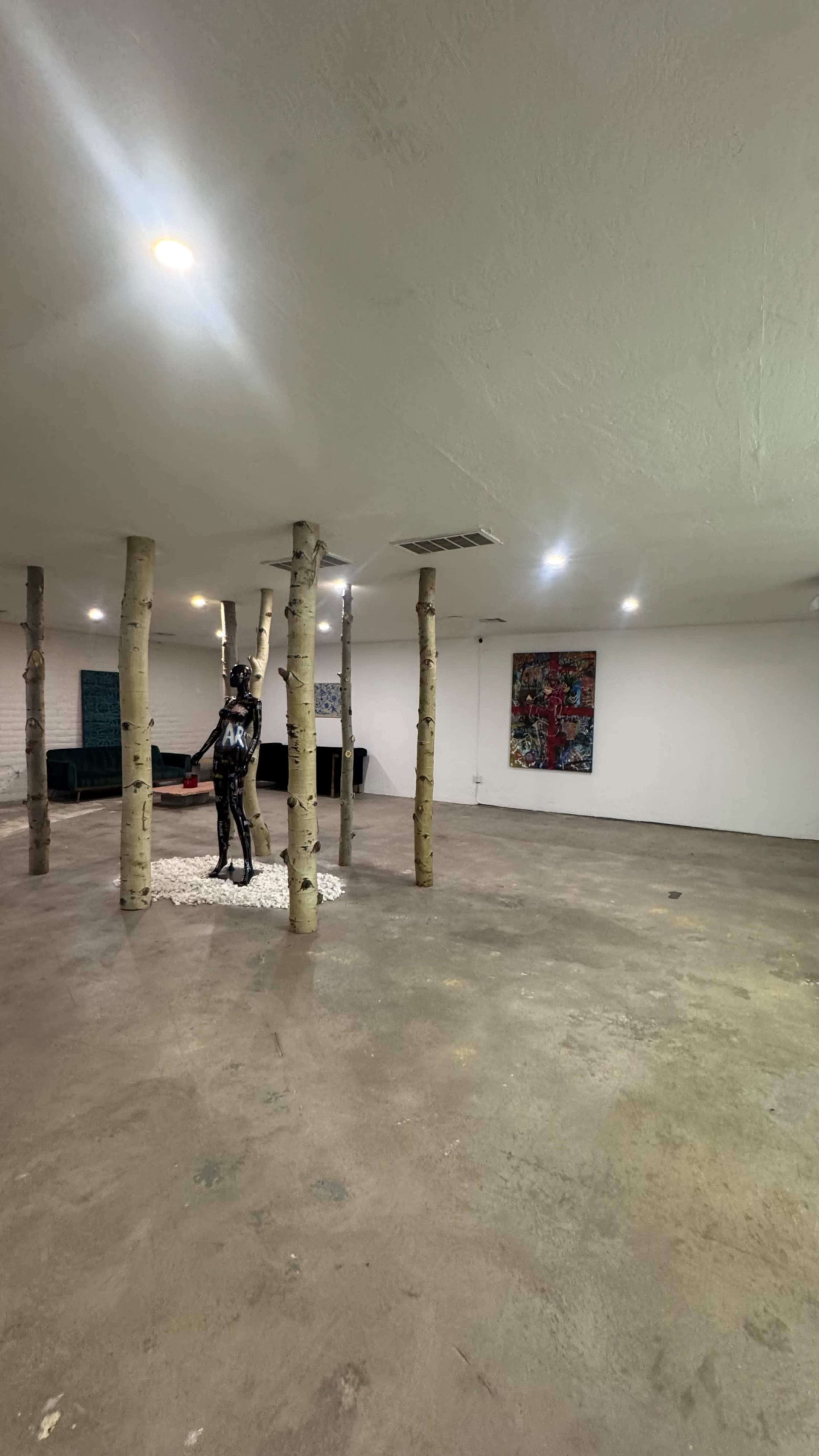 The image shows a spacious, empty room featuring several vertical wooden pillars and a sculpture in the center, along with art pieces on the walls.