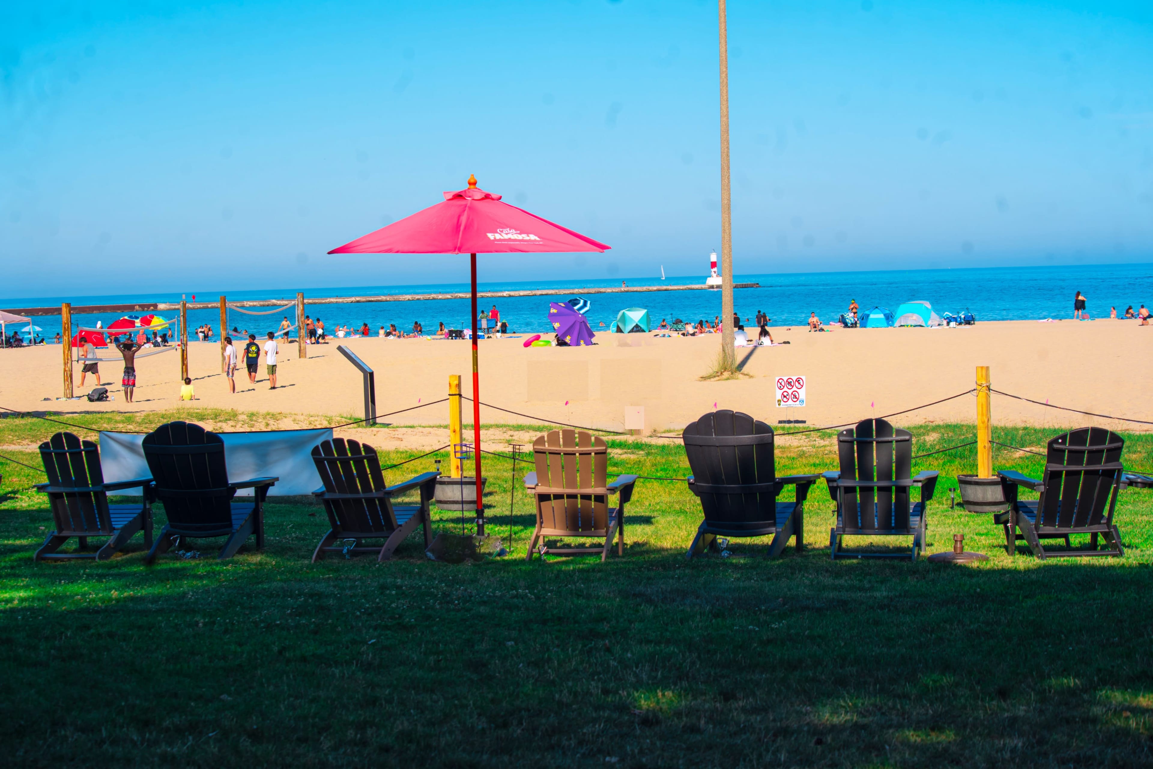 The image shows several wooden lounge chairs aligned on a grassy area overlooking a sandy beach with umbrellas and beachgoers in the distance.