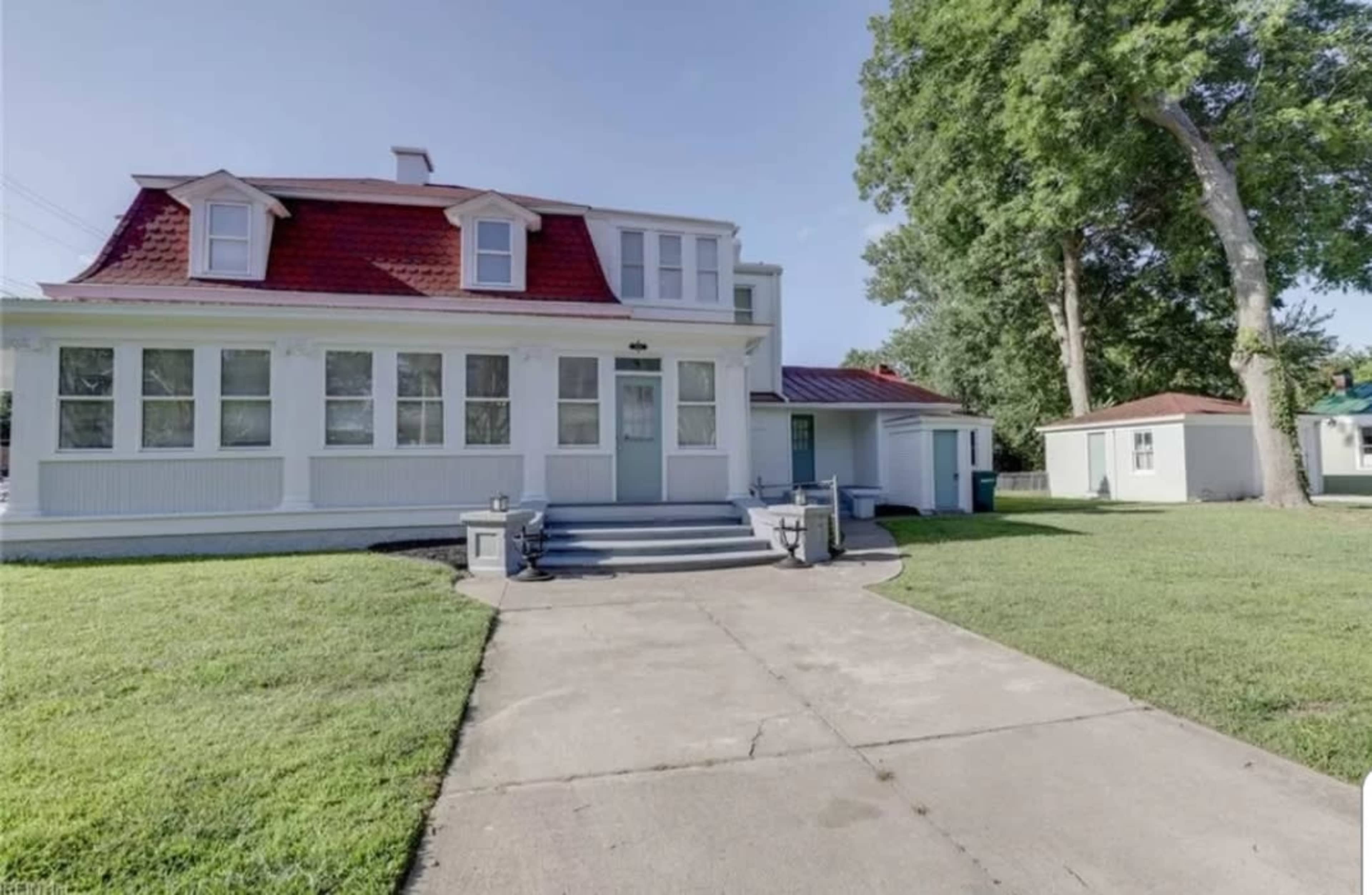 A two-story house with a red roof, white siding, and a concrete pathway leading to the entrance, surrounded by a lawn and trees.