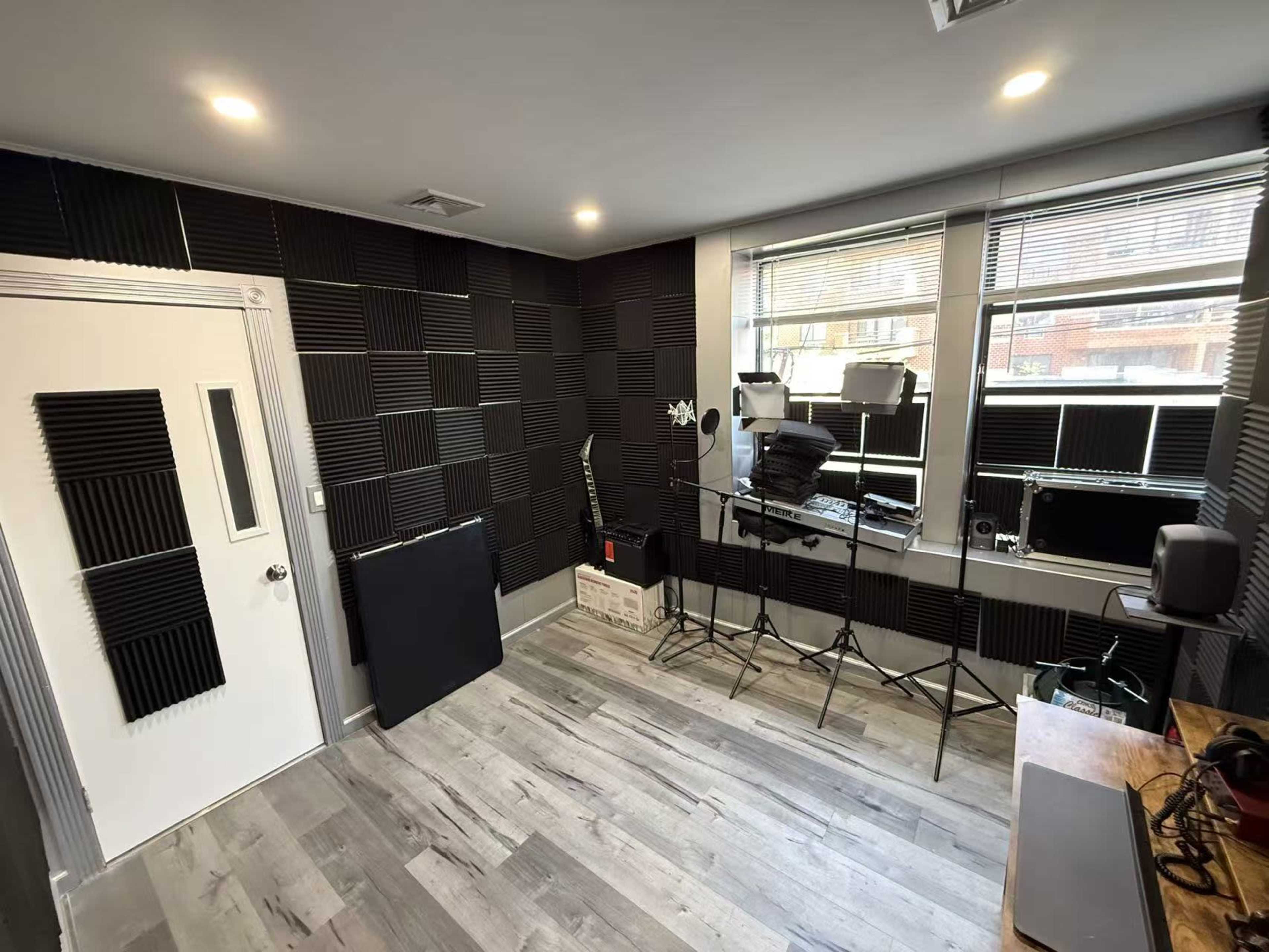 The image shows a soundproofed room with black acoustic foam panels on the walls, wooden flooring, and equipment stands near large windows.