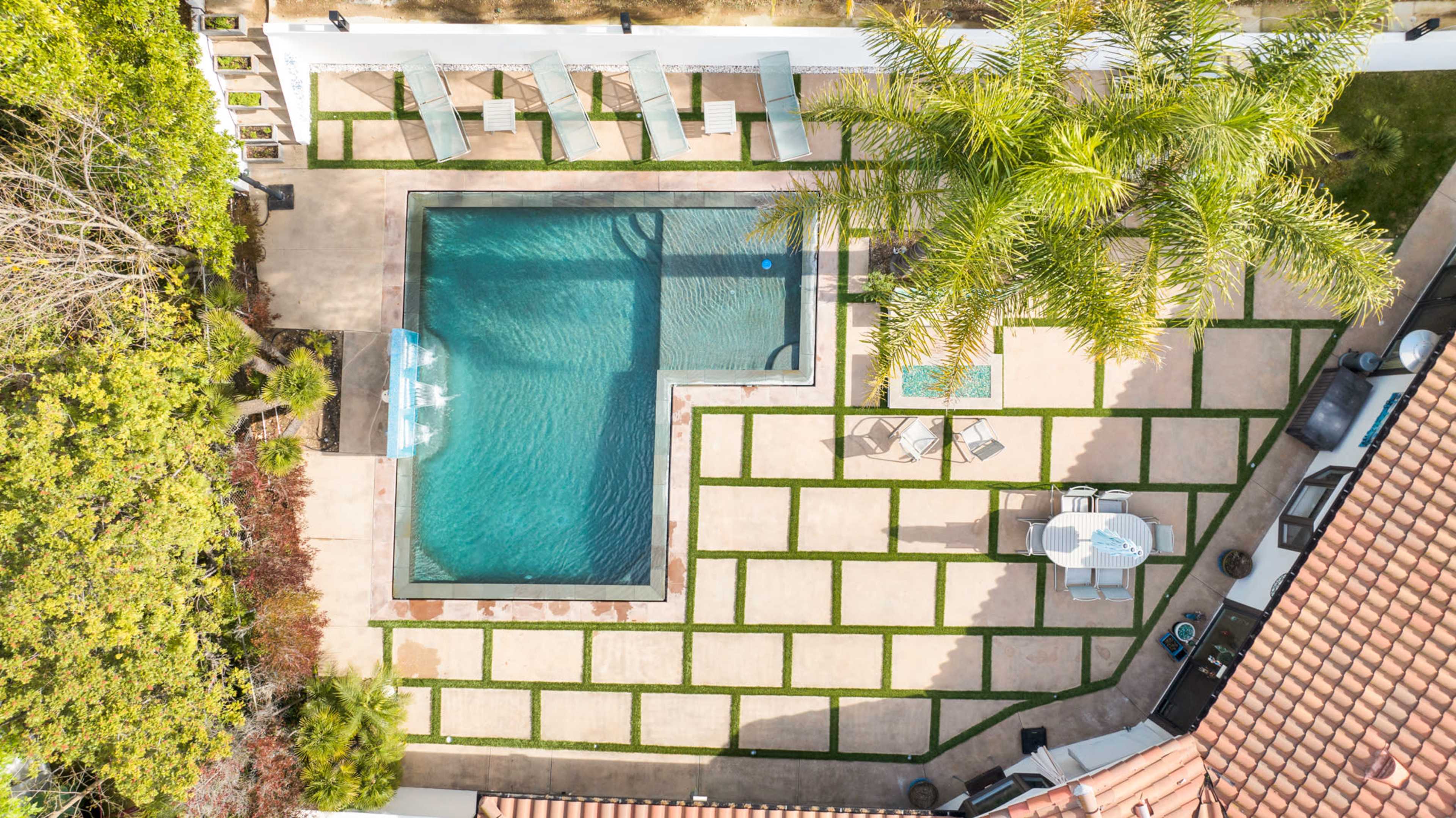 An aerial view shows a landscaped backyard with a rectangular swimming pool surrounded by lounge chairs and a dining area.
