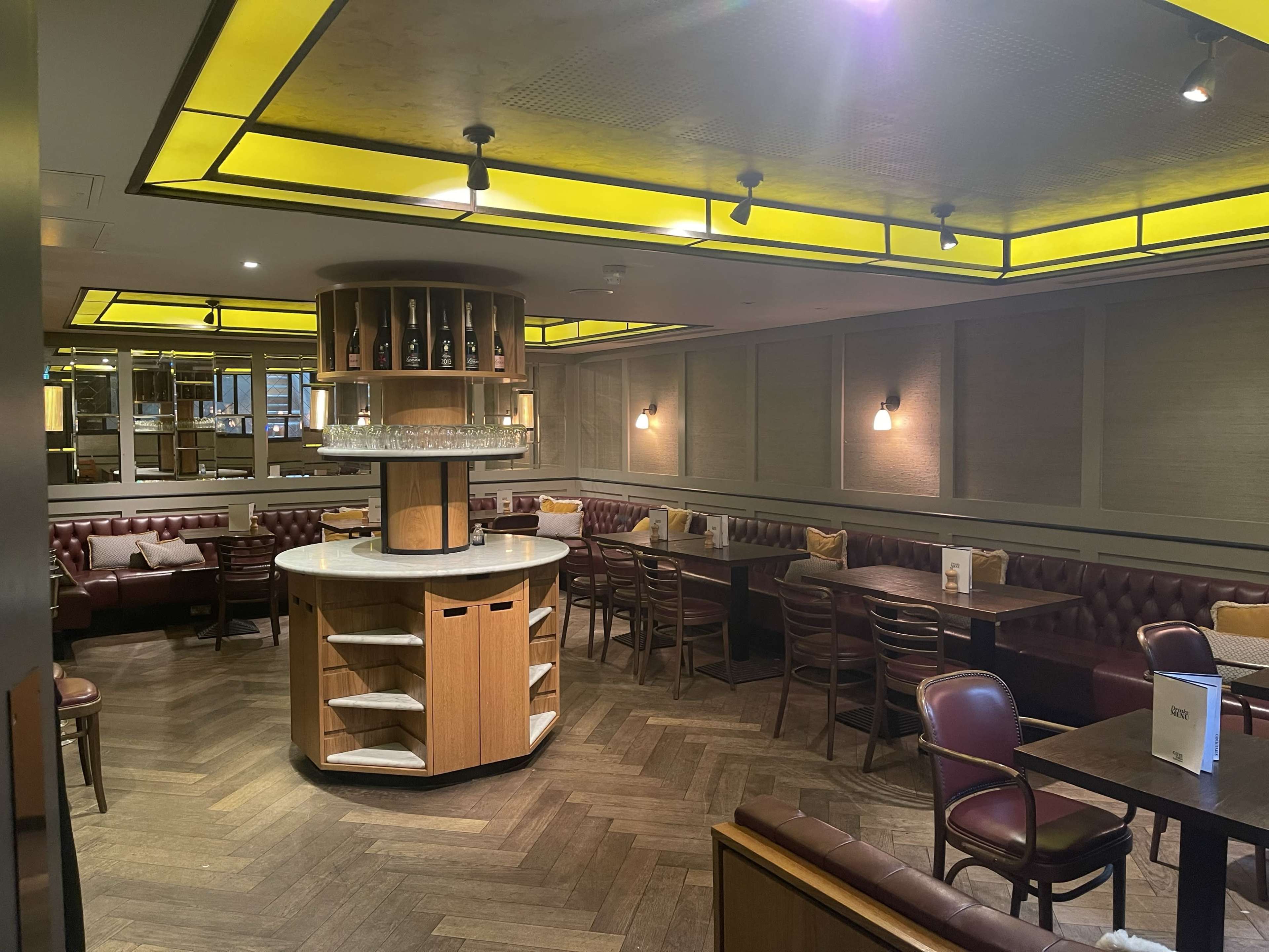 The image shows an upscale restaurant interior featuring wooden furniture, a central circular bar with shelves, and cozy booth seating along the walls.