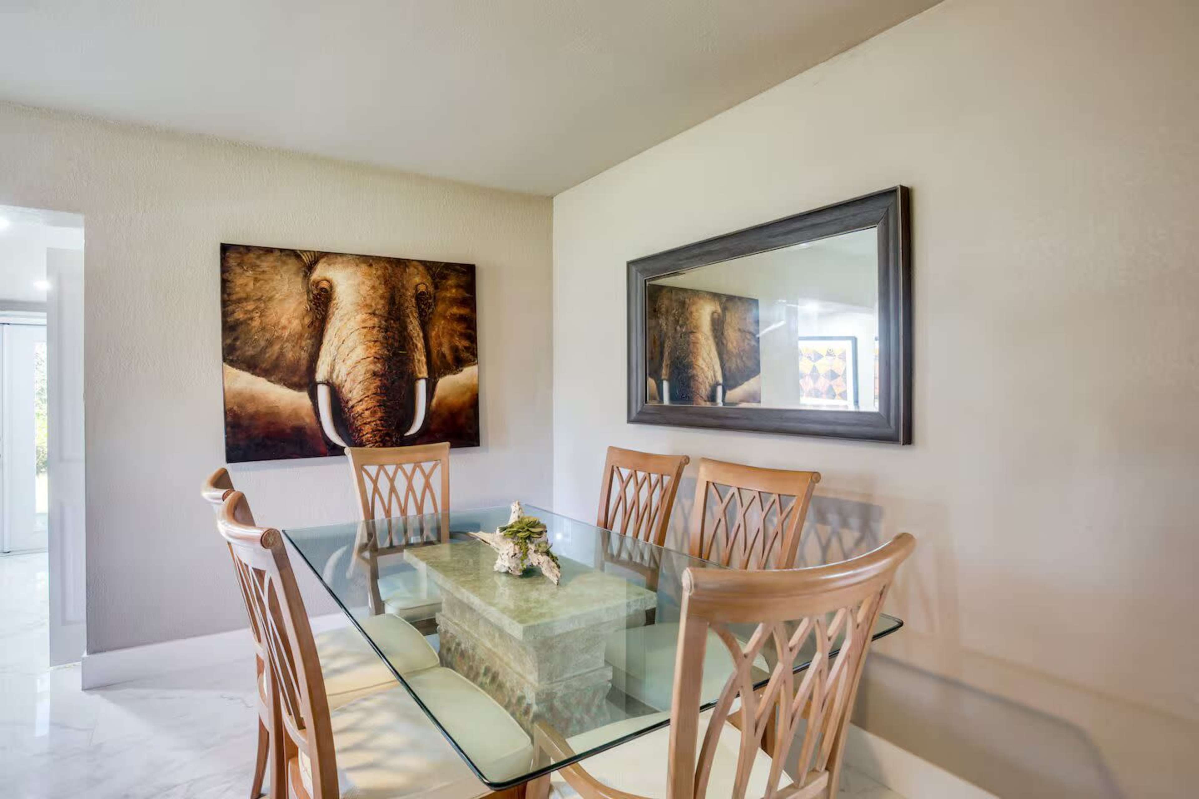 A glass dining table with six wooden chairs is positioned in a room featuring an elephant painting and a mirror on the wall.