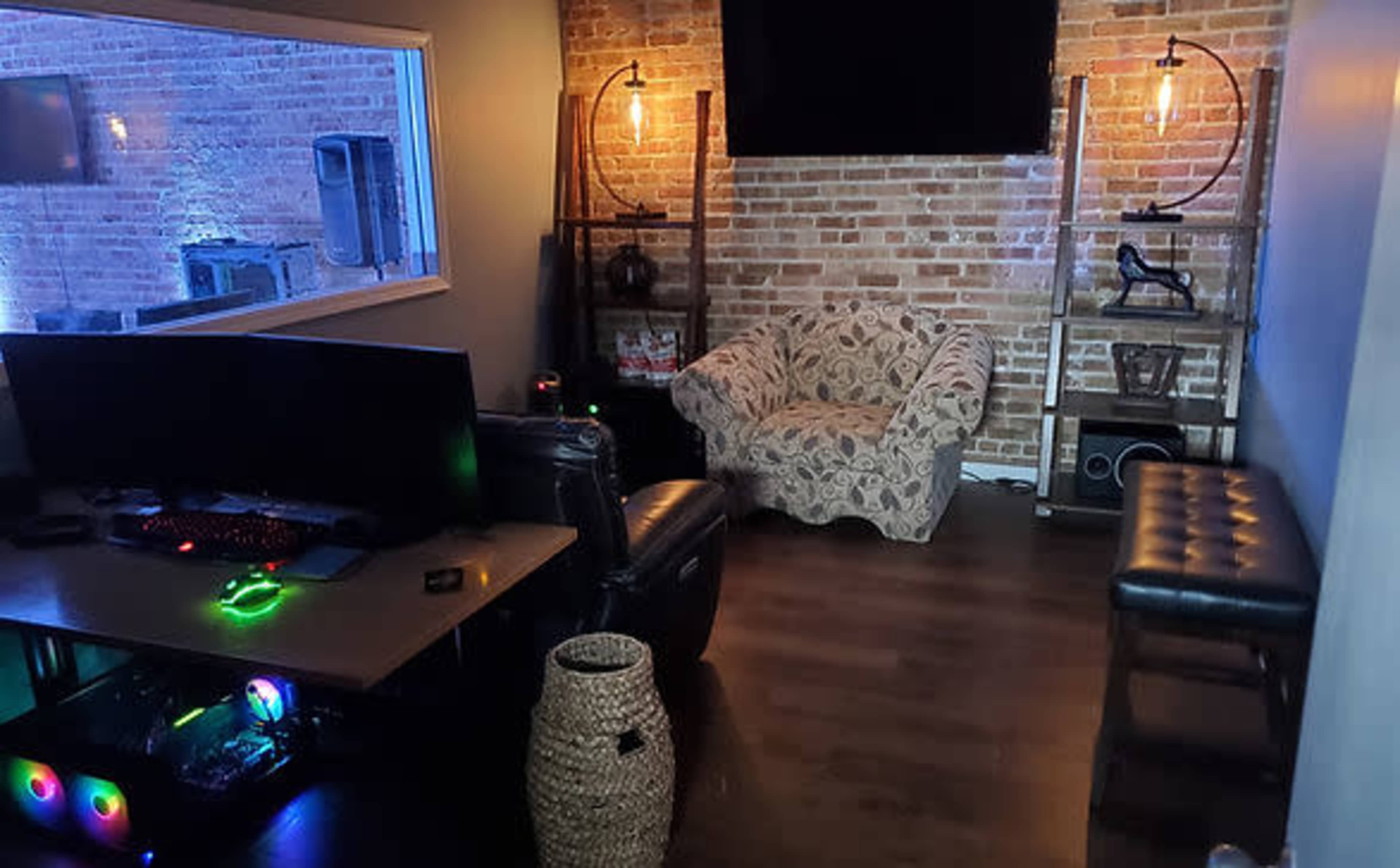 A cozy room with a sectional couch, a large computer setup with colorful lighting, and exposed brick walls.