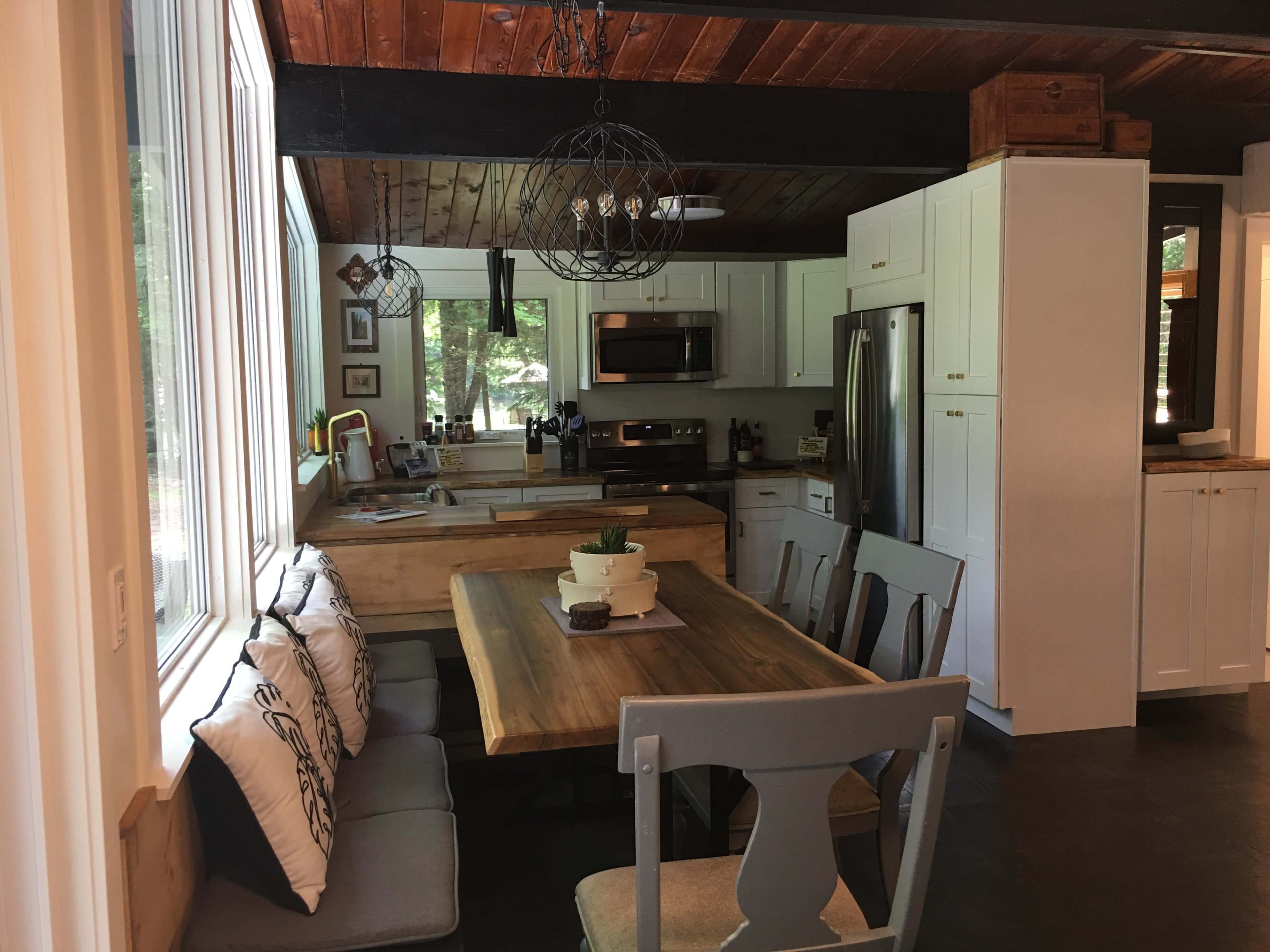 A modern kitchen with a wooden dining table, bench seating, and white cabinetry, illuminated by natural light from large windows.