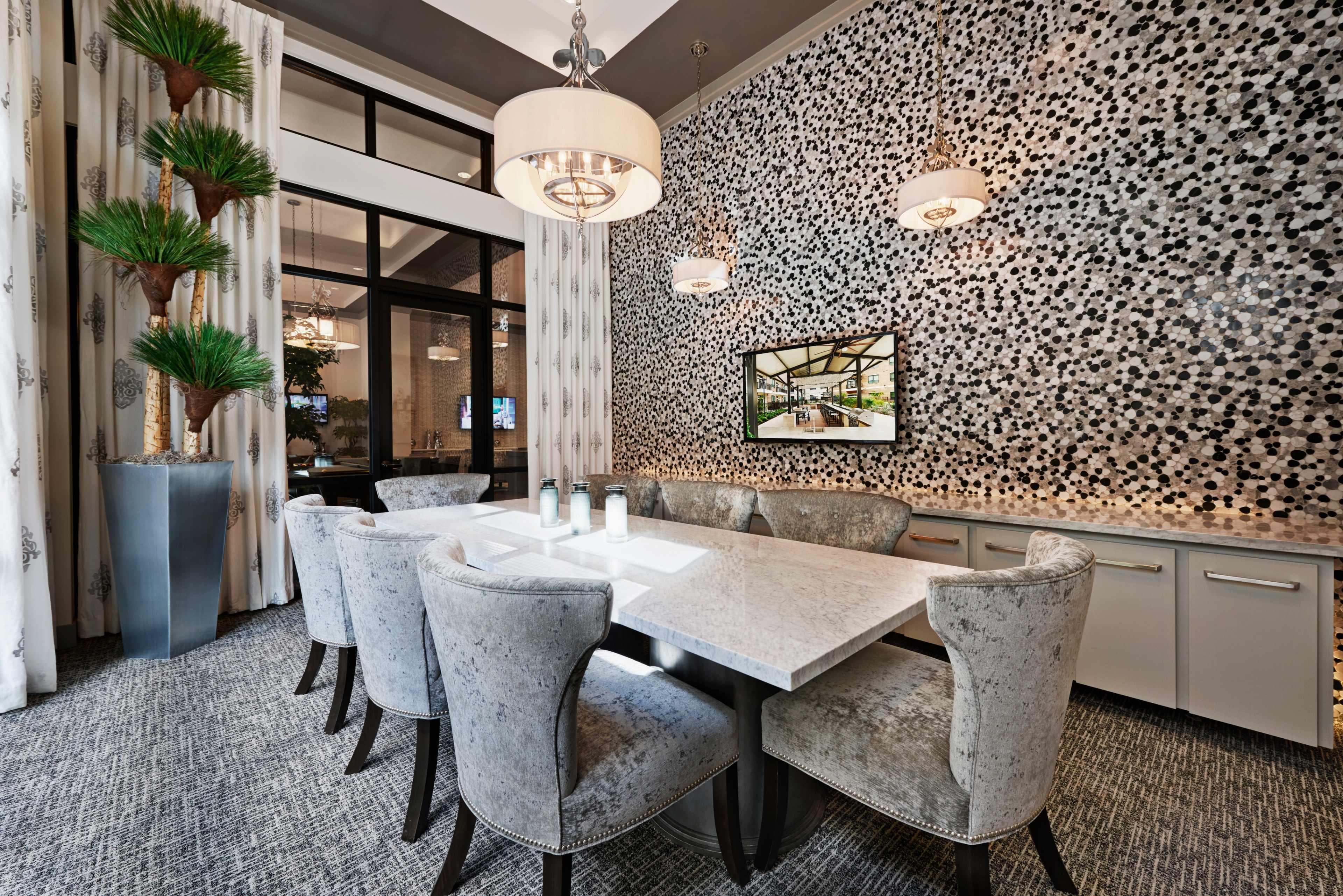 The image shows a modern dining area with a large marble table surrounded by upholstered chairs, a decorative wall featuring black and white tiles, and a tall planter with green plants.