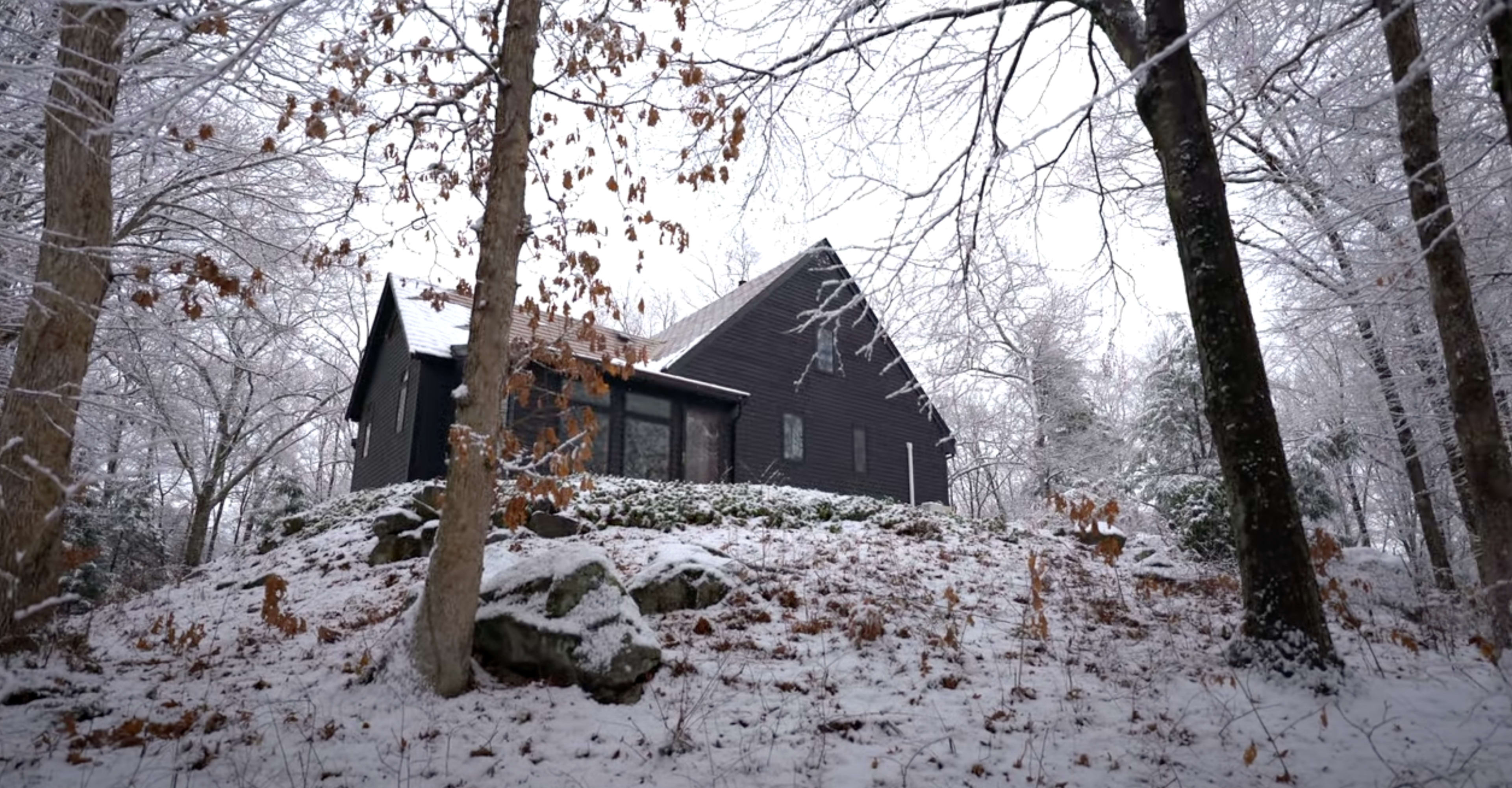 A modern black house with a sloped roof is surrounded by snow-covered trees and a rocky terrain.