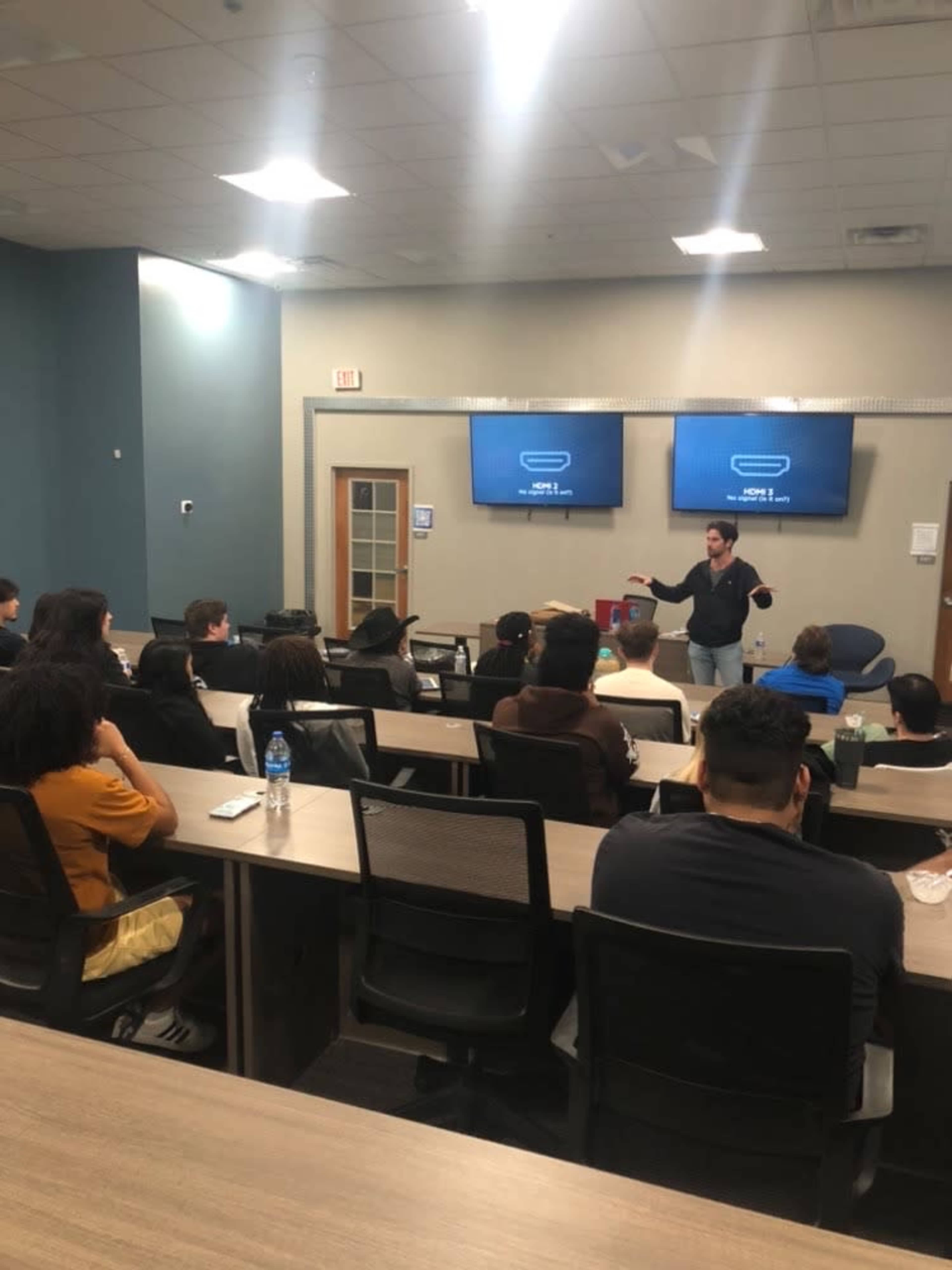 A speaker addresses a seated audience in a classroom equipped with two large screens displaying information.