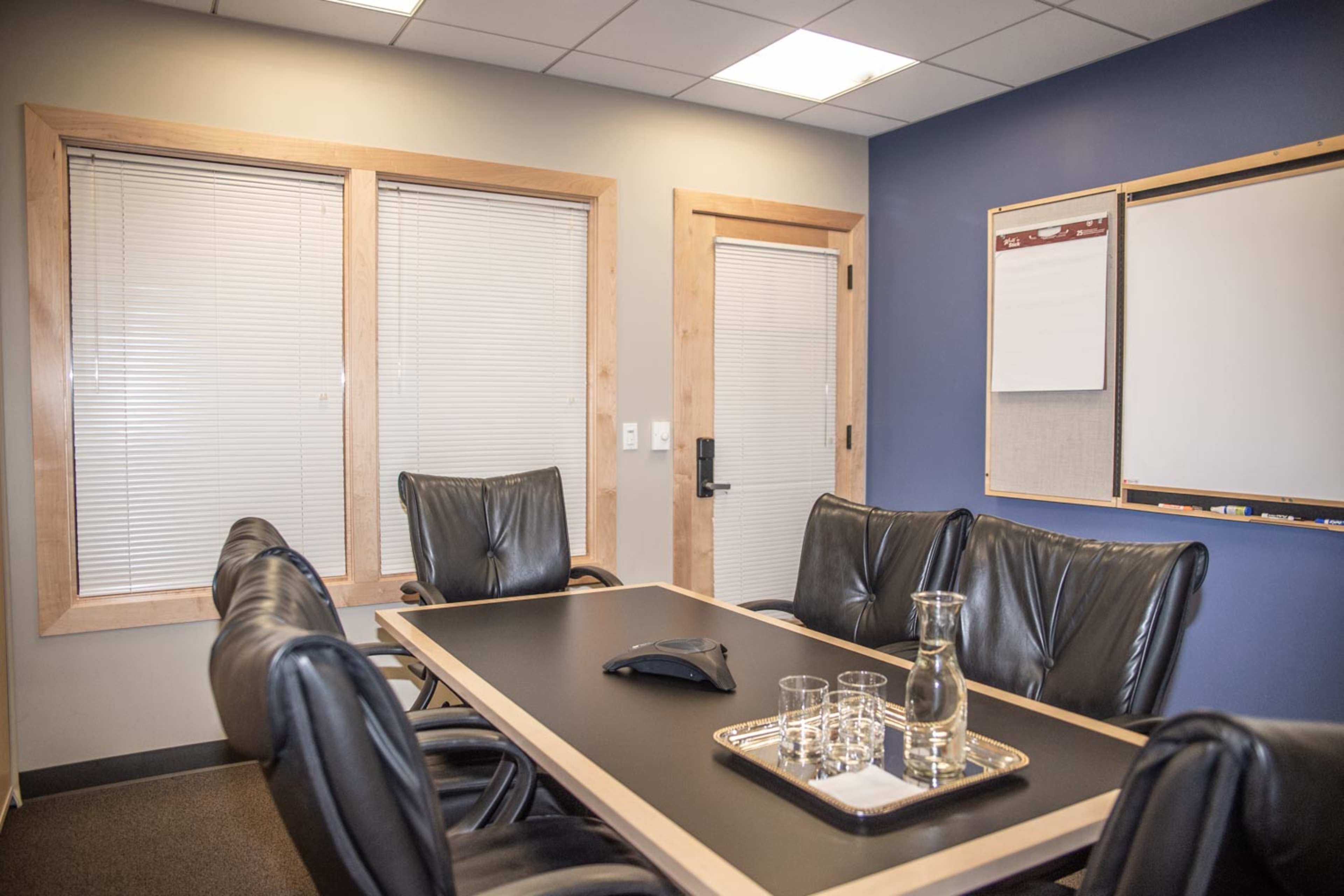 The image depicts a small conference room featuring a long table surrounded by black leather chairs, a glass pitcher, and a few drinking glasses, with a whiteboard and a door in the background.