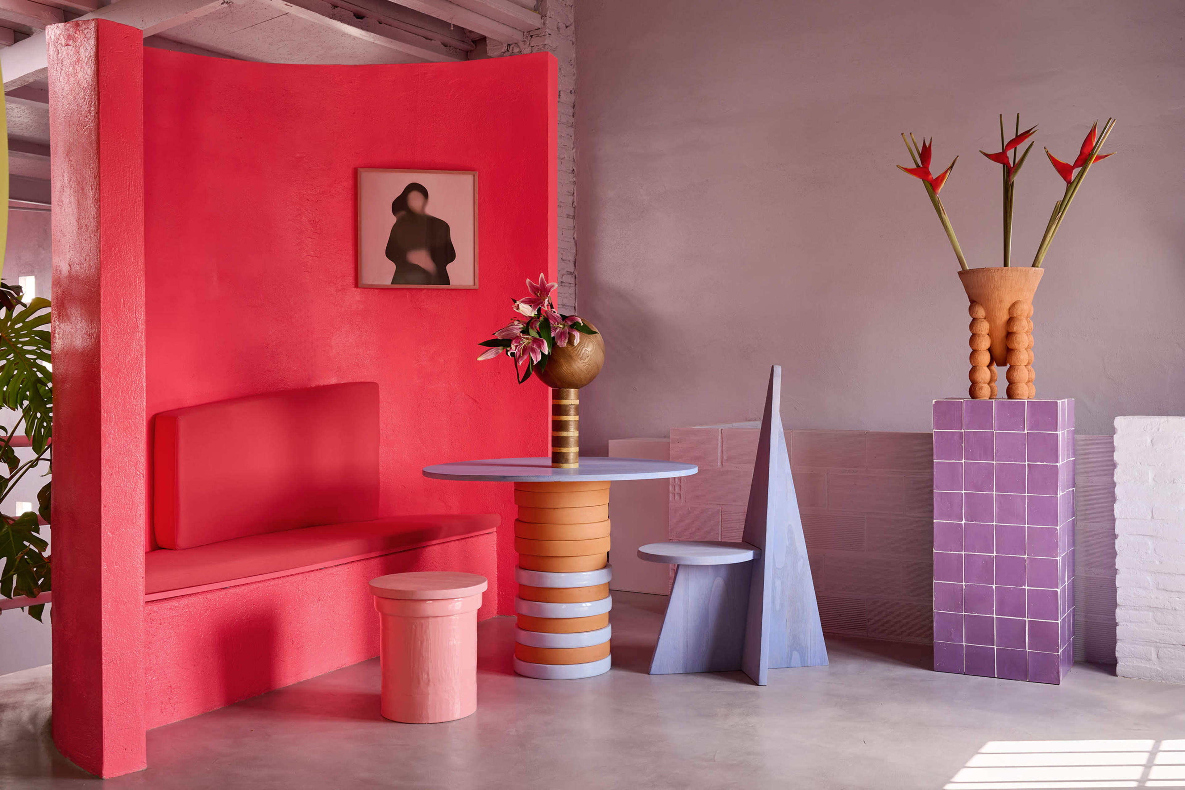 The image features a modern interior design setup with colorful furniture, including a pink bench, a round table with striped legs, and a geometric pedestal, accompanied by floral arrangements and a framed artwork on the wall.