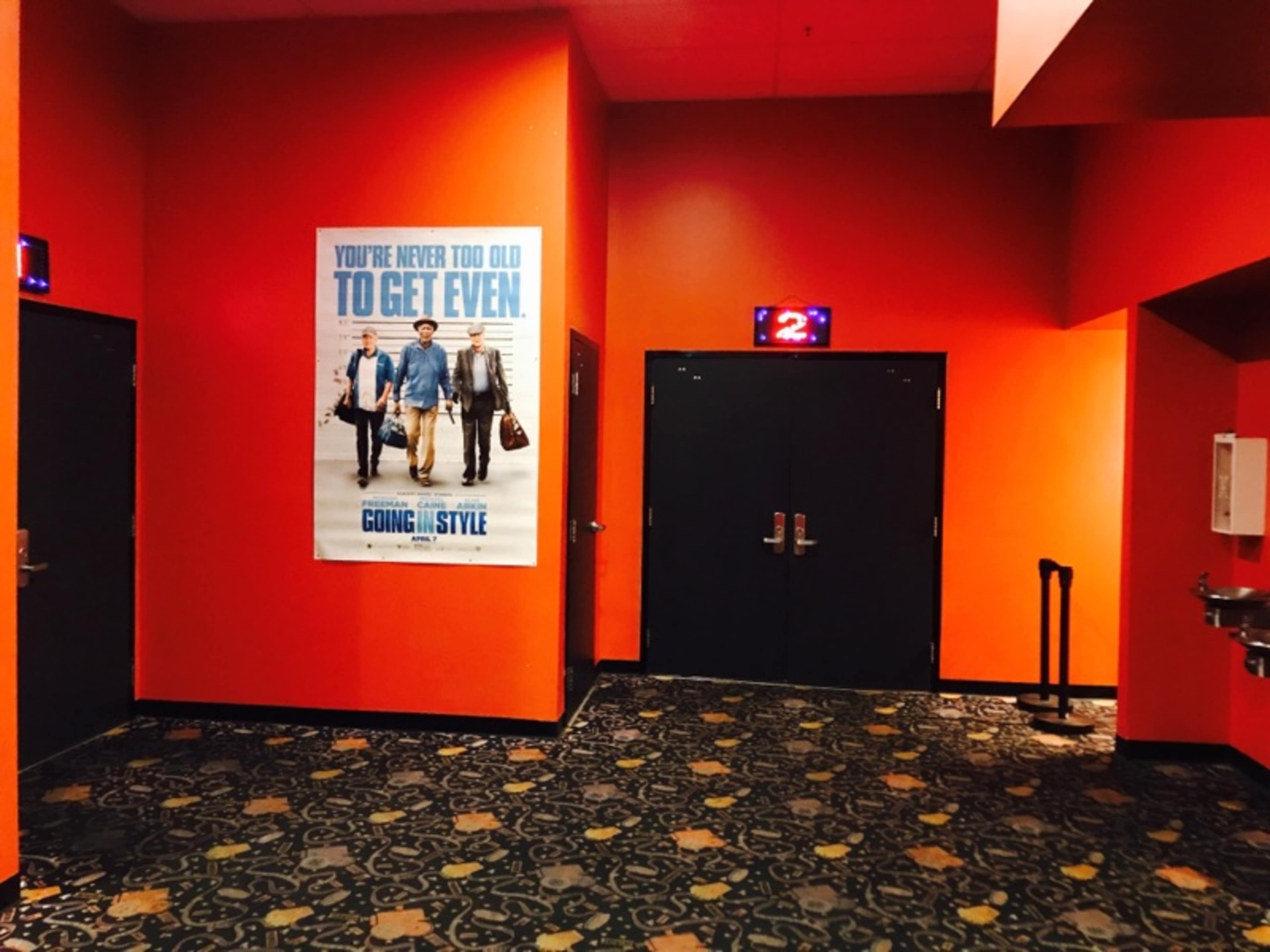 A brightly colored movie theater corridor with a large poster for "Going in Style" and two closed doors marked with the number 2.