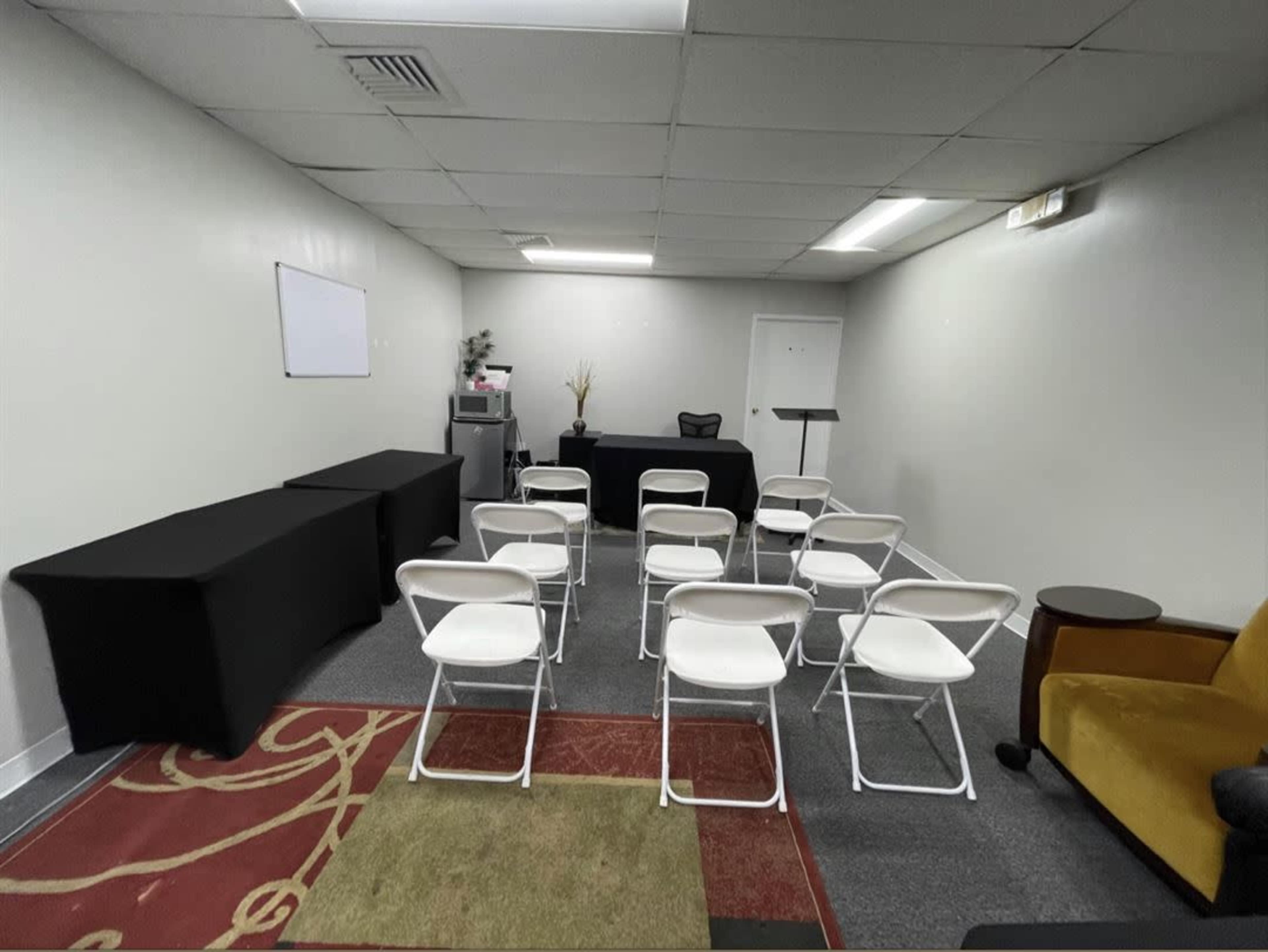 A small, sparsely furnished meeting room with several white folding chairs facing a black table and a podium.