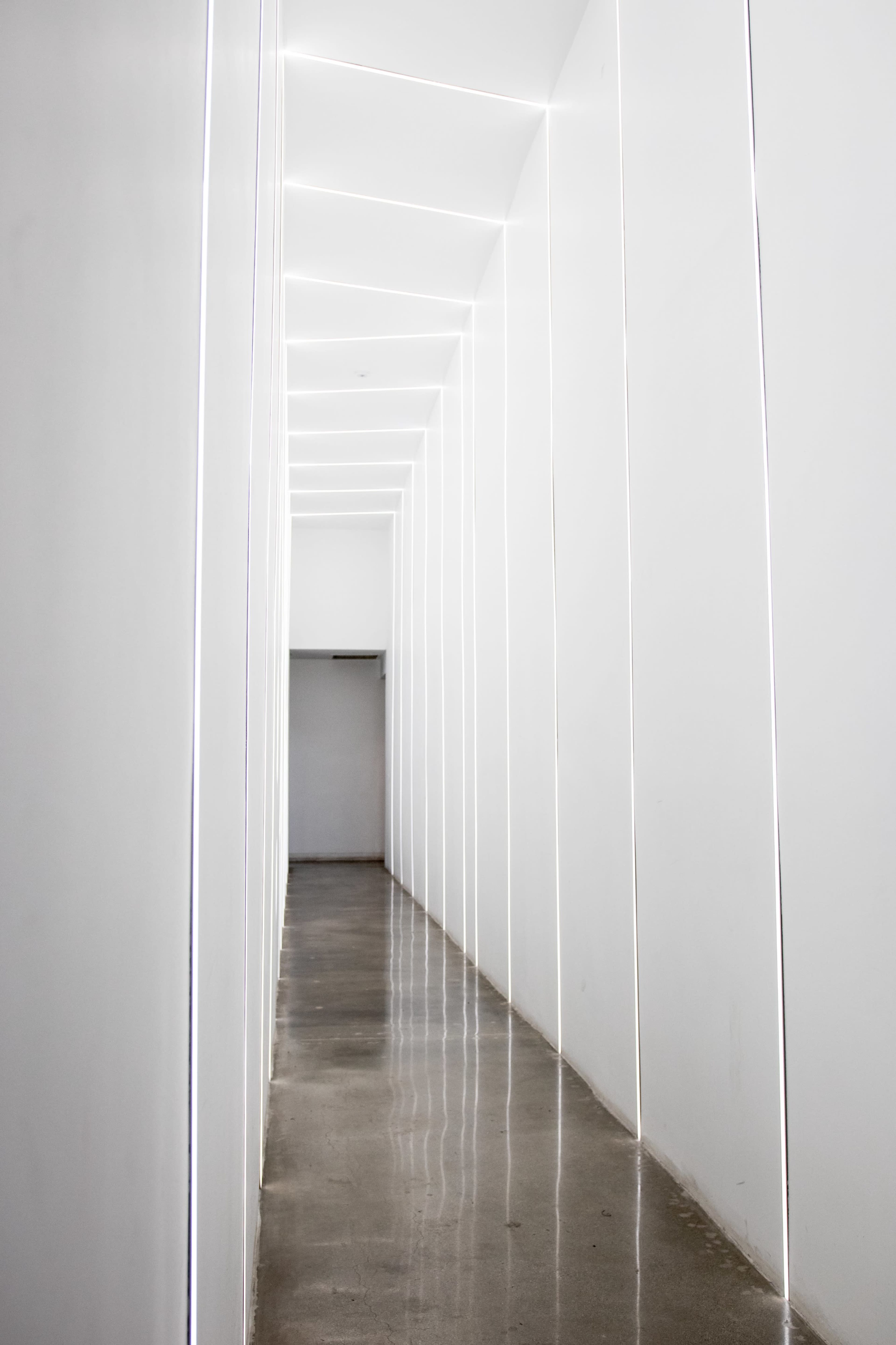 A long, narrow hallway features white walls and lighting strips that run along the edges, reflecting on a polished concrete floor.