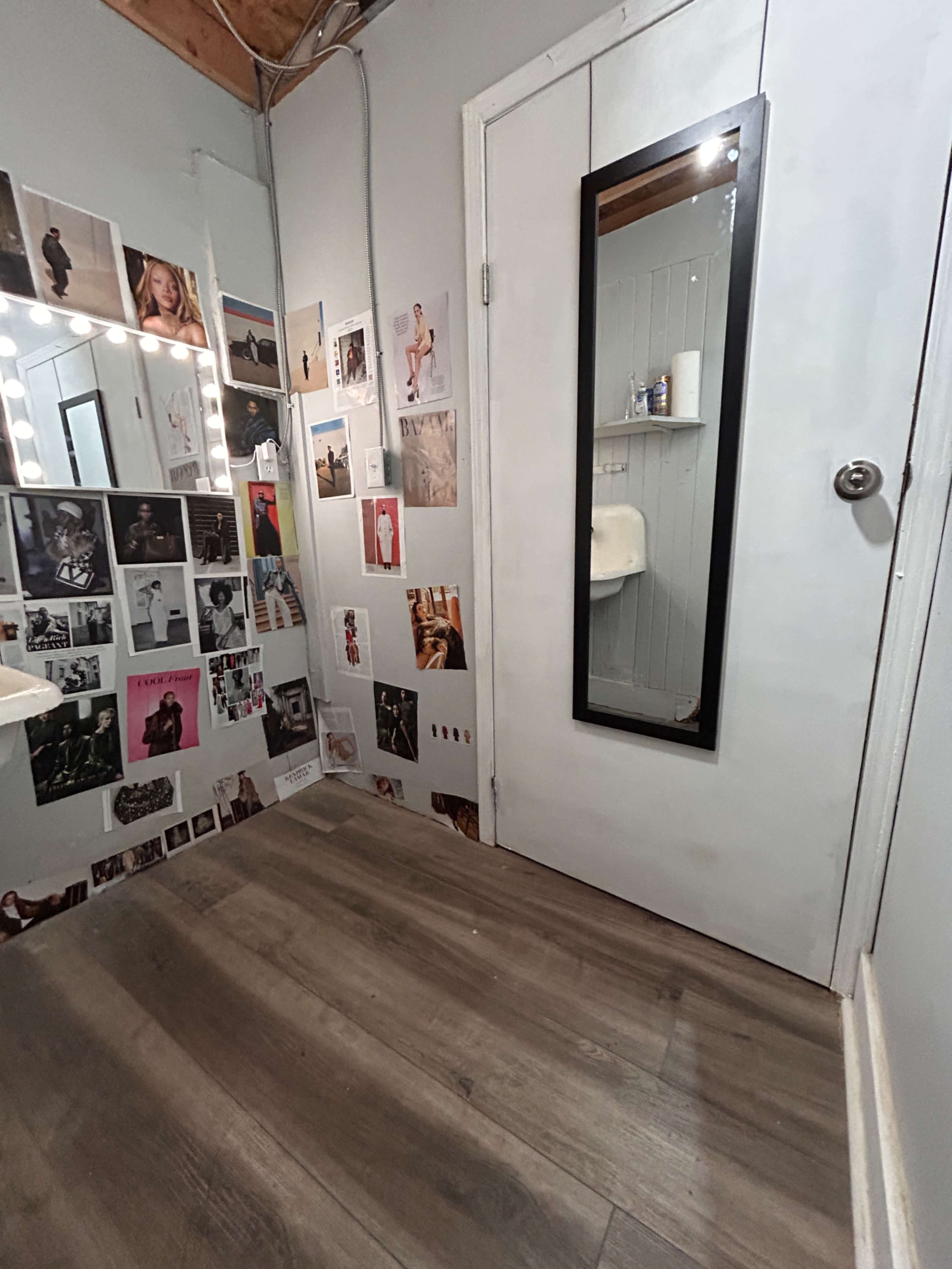 The image shows a room with a mirror on the door and walls covered in various photographs, next to a lighted vanity mirror and a small sink.