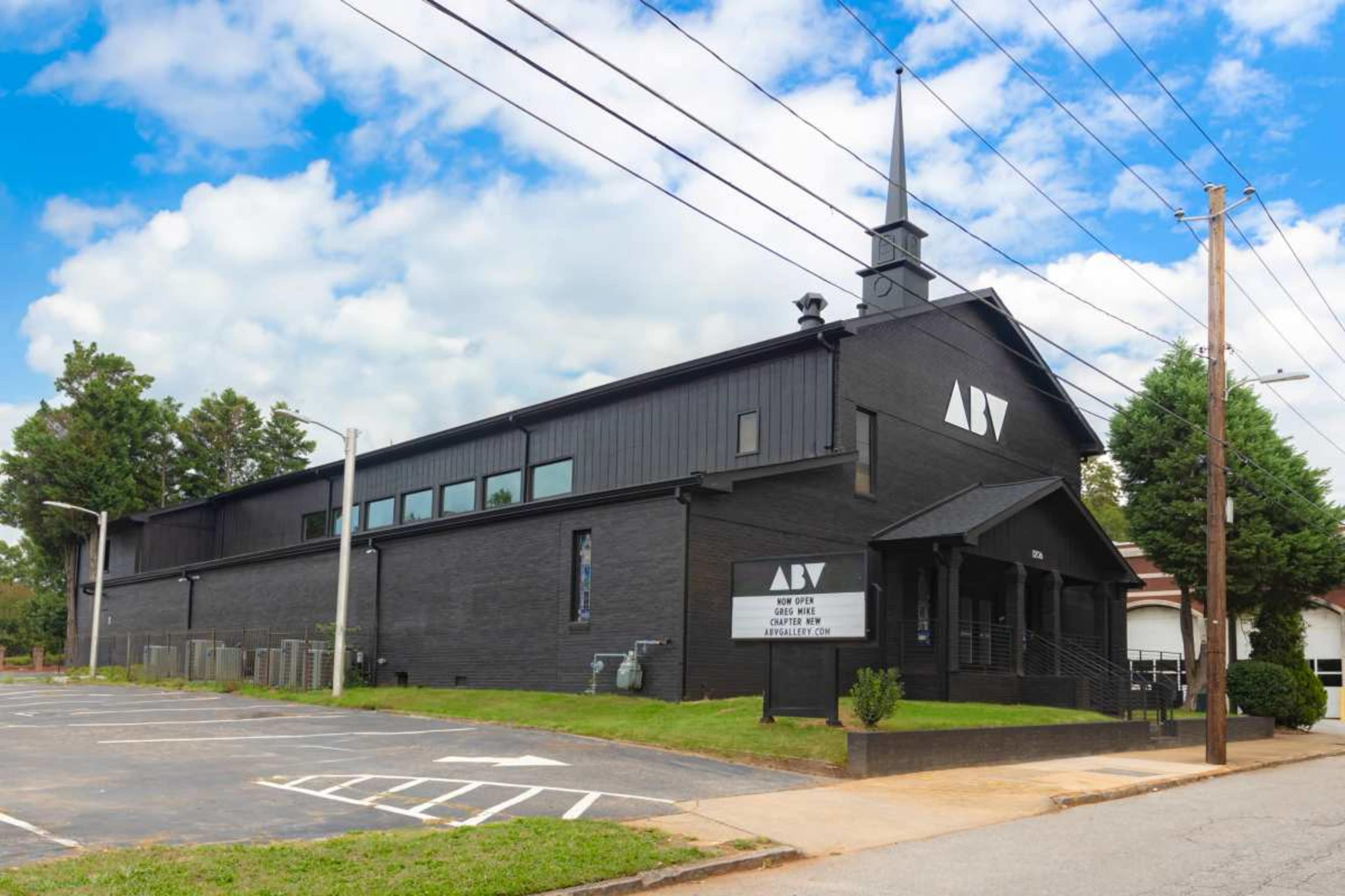 The image shows a modern black building with a steeple surrounded by trees and a partially empty parking lot under a cloudy sky.