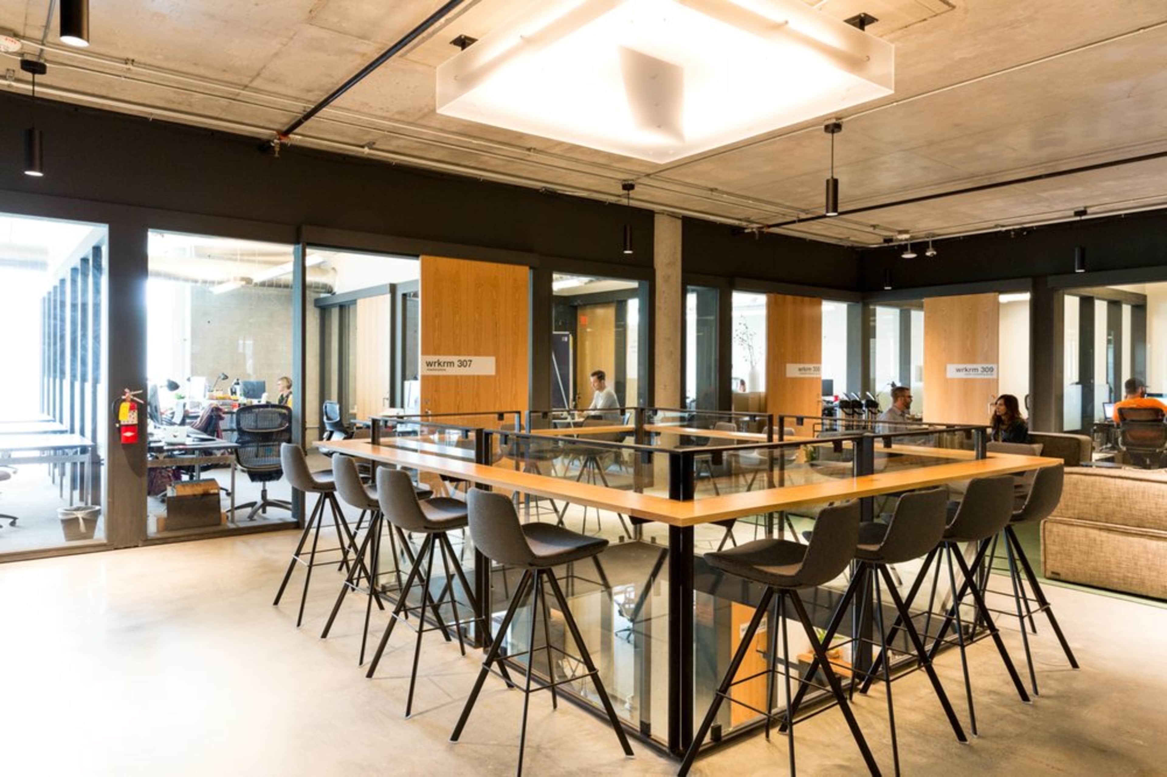 A modern office space featuring a large glass-topped table surrounded by high stools, with contemporary workstations visible in the background.