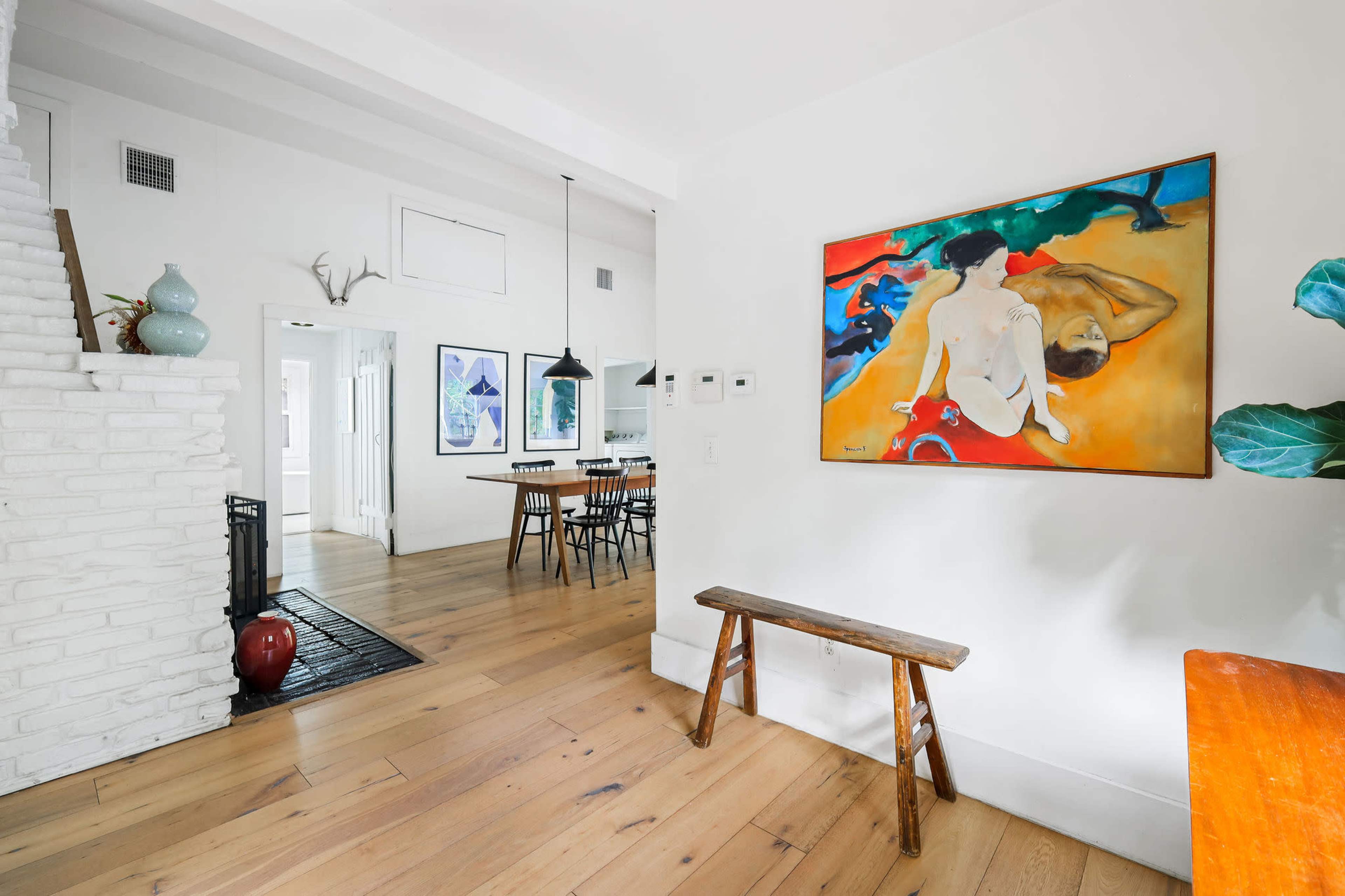 A hallway features a wooden bench, a colorful painting on the wall, and a view of a dining area with a long table and chairs.