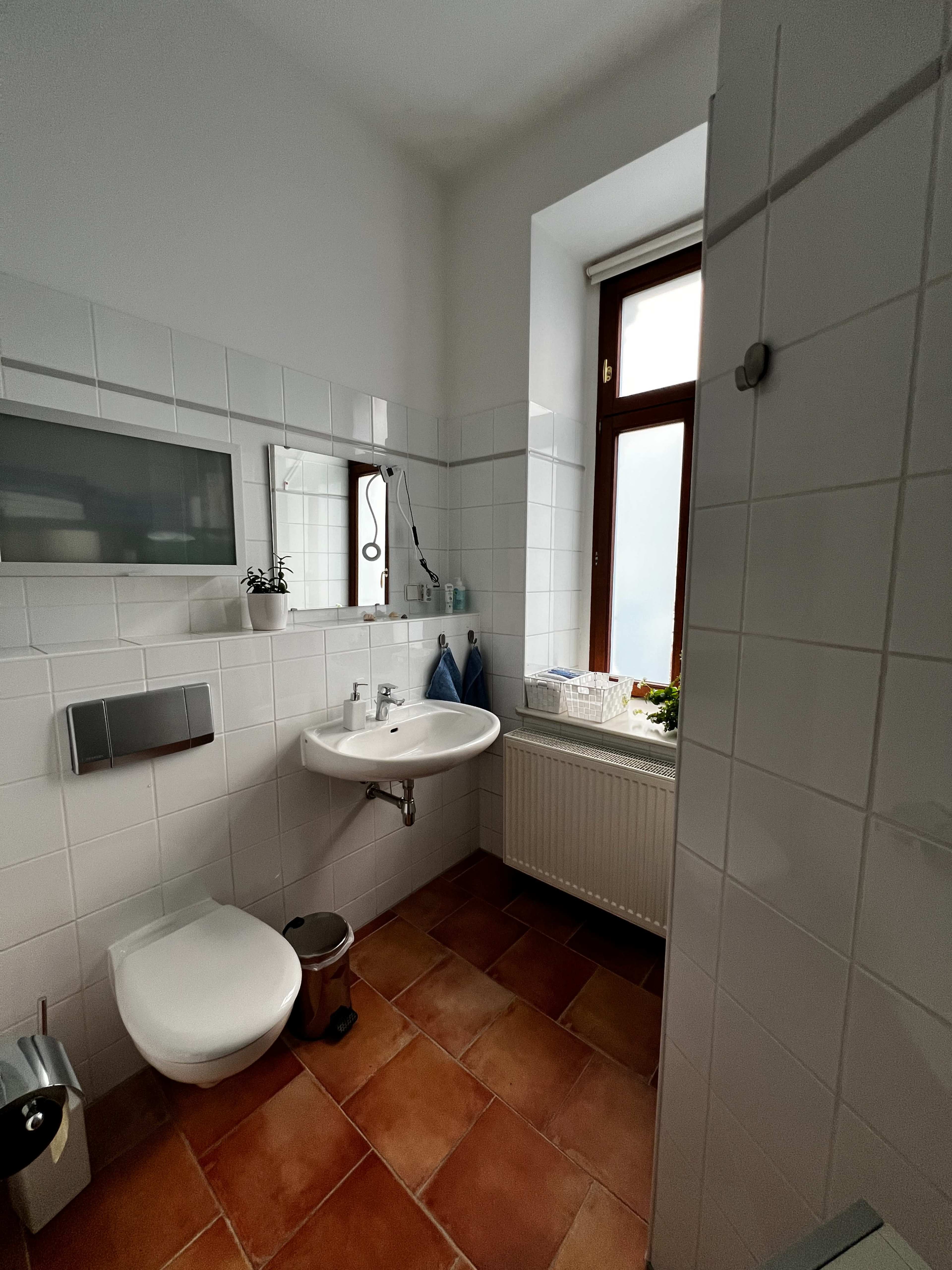 The image shows a tiled bathroom featuring a toilet, sink, mirror, and a window with light streaming in.