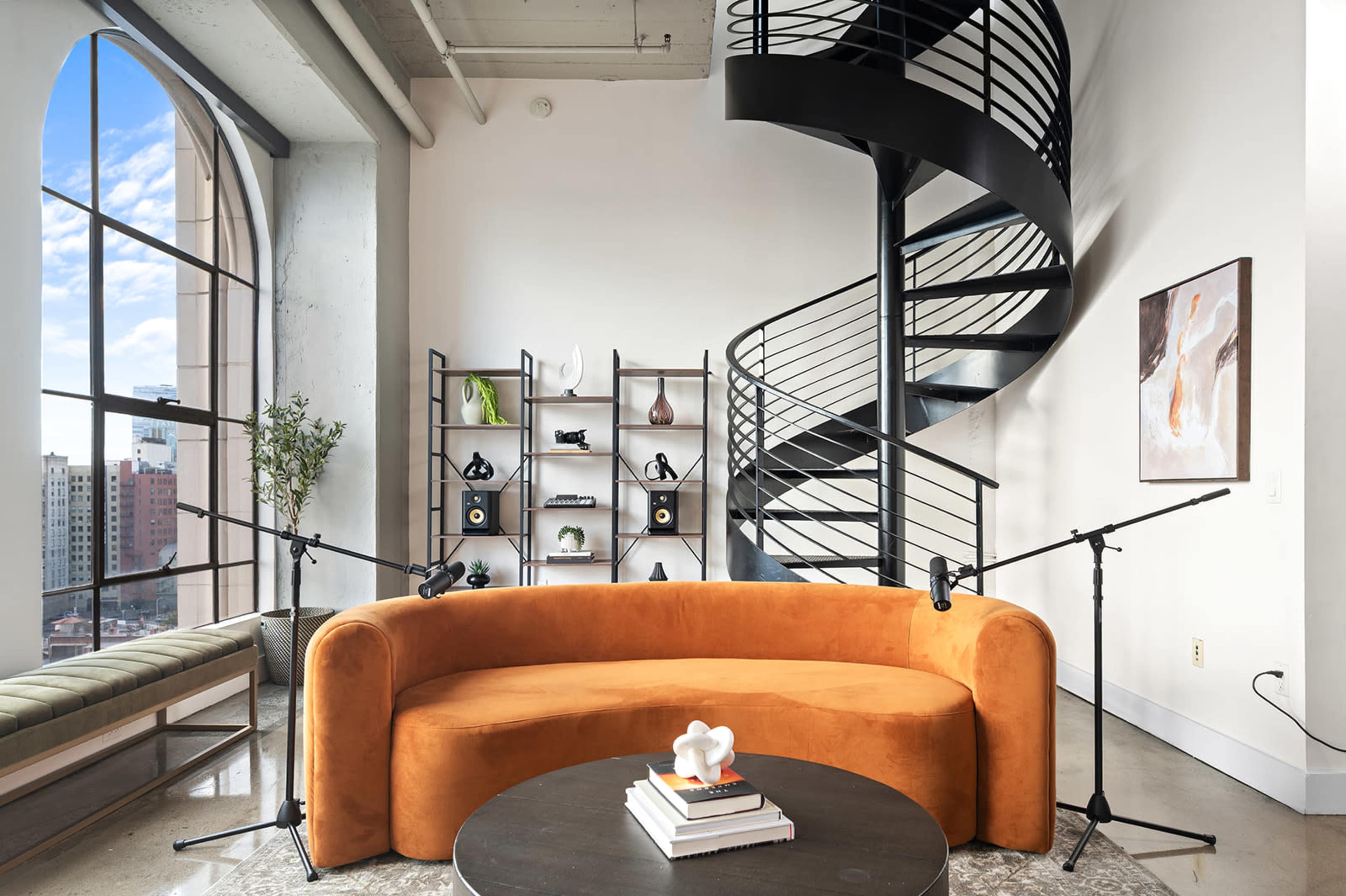 The image shows a modern living space featuring a rounded orange couch, a black spiral staircase, and a shelving unit displaying various decorative items.