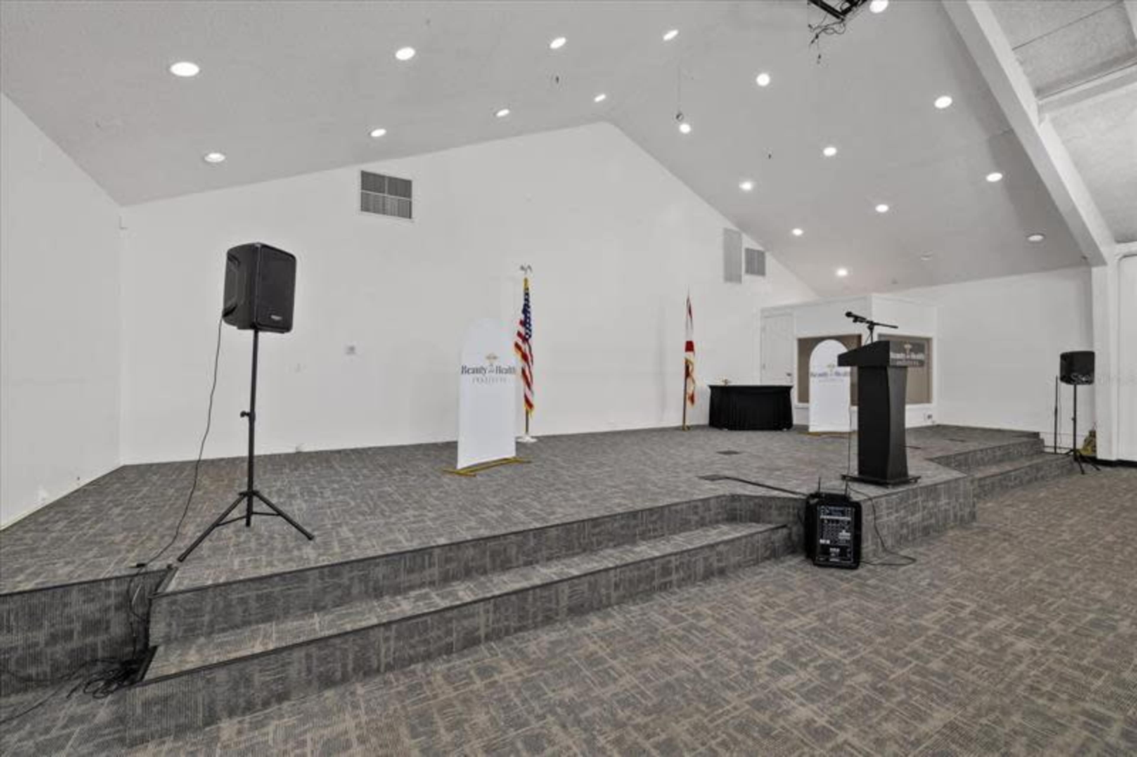 The image shows a spacious indoor venue with a raised stage, featuring equipped sound systems, a podium, and flags at the front.