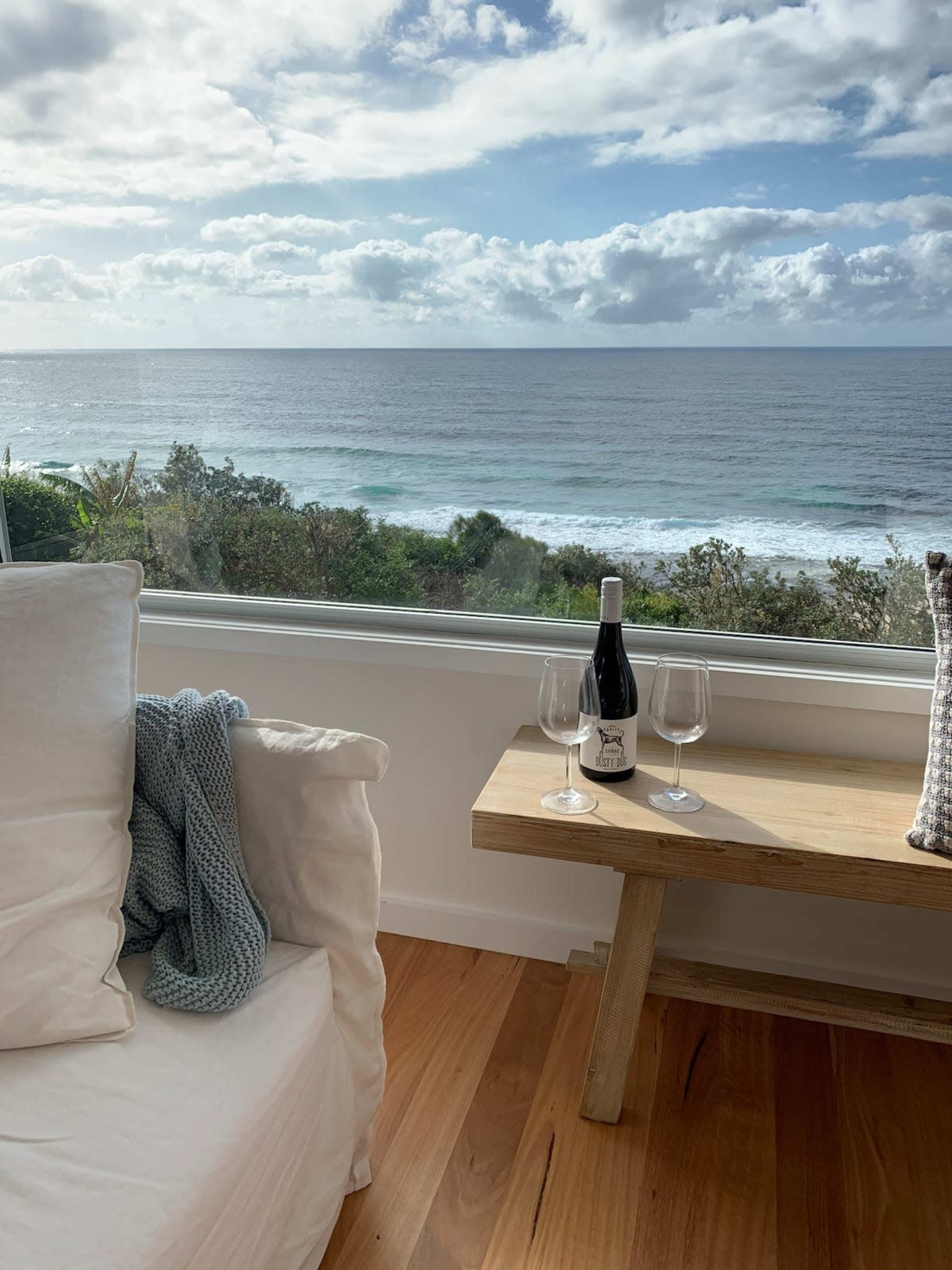 A cozy interior features a light-colored sofa next to a wooden table with a bottle of wine and two glasses, overlooking a calm ocean and cloudy sky through a large window.
