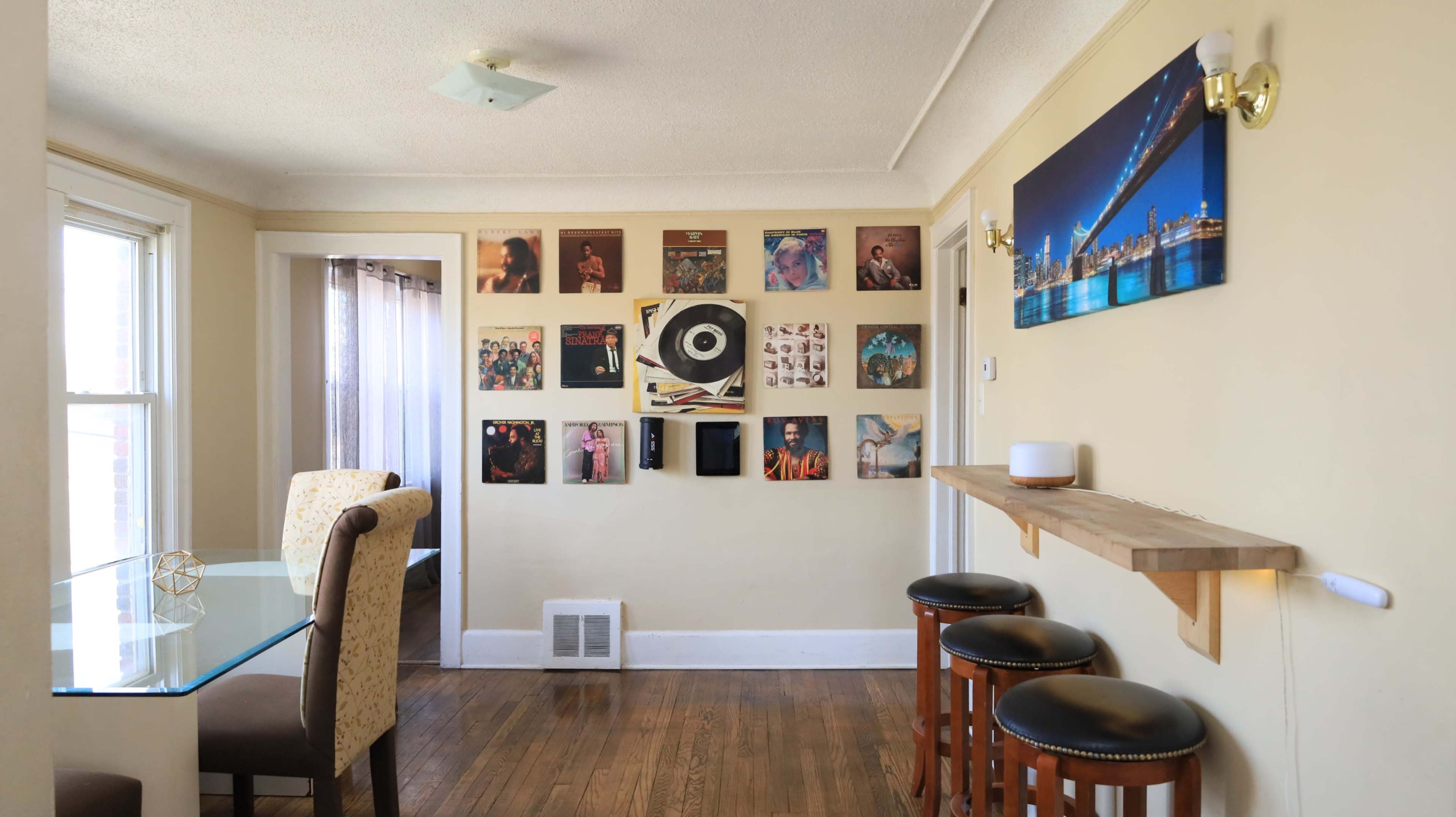 A dining area features a glass table and three wooden bar stools, with a wall adorned with framed album covers and a skyline photo.