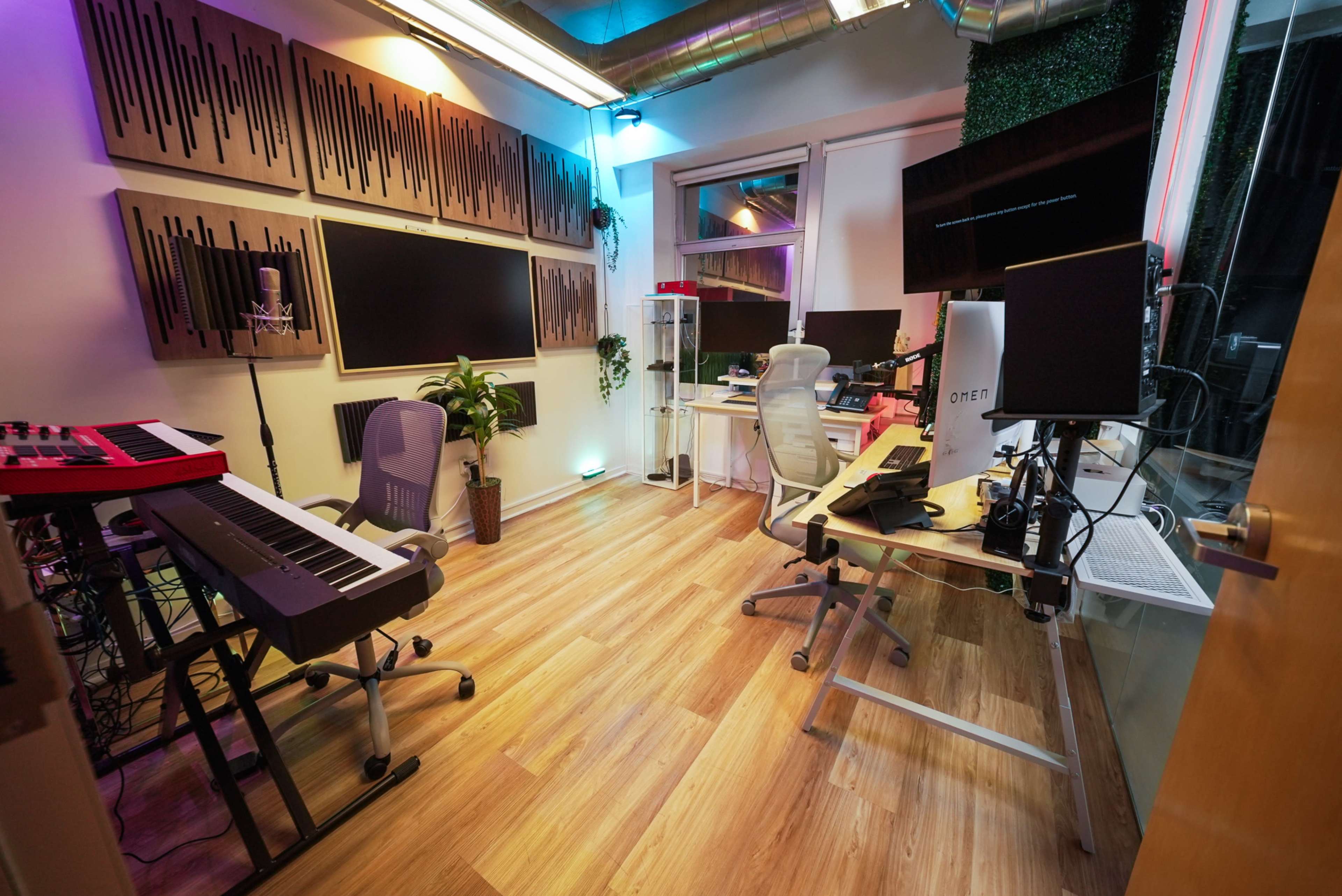The image shows a modern recording studio with sound panels on the walls, multiple computer monitors, a keyboard, and ample workspaces including chairs and plants.