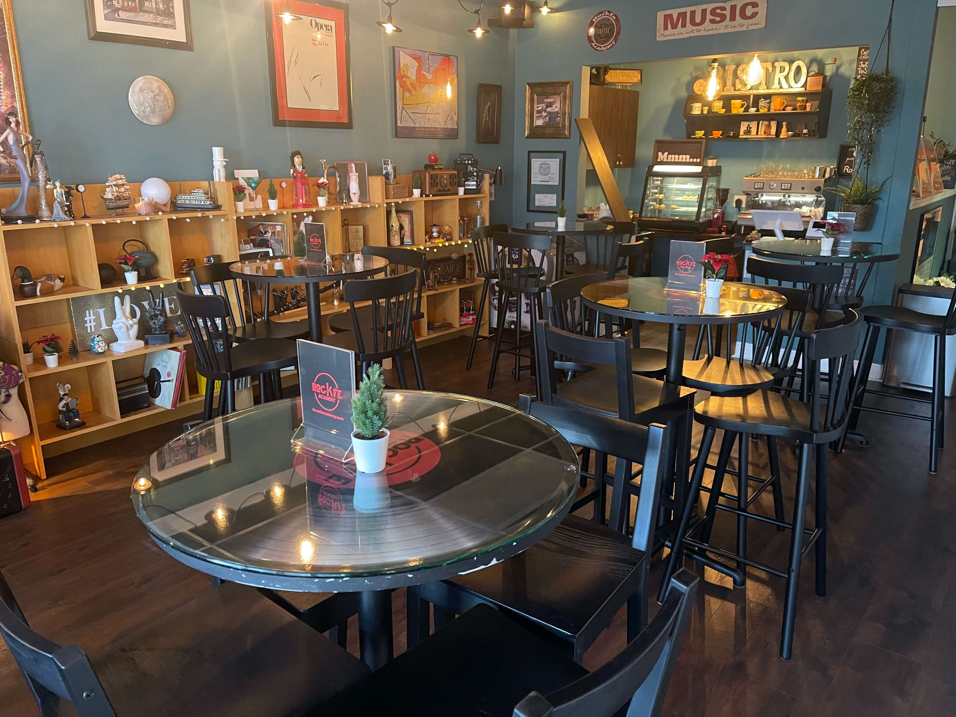 The image shows a cozy café interior with wooden shelves displaying various decor items, black tables, and chairs arranged around the space.